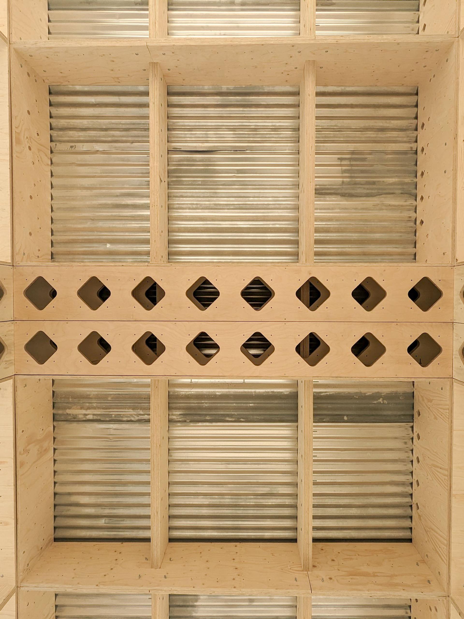Ceiling with a grid of light spruce beams and corrugated metal panels, featuring a central wooden section with diagonal square cut-outs in two rows—an elegant example of timber construction.