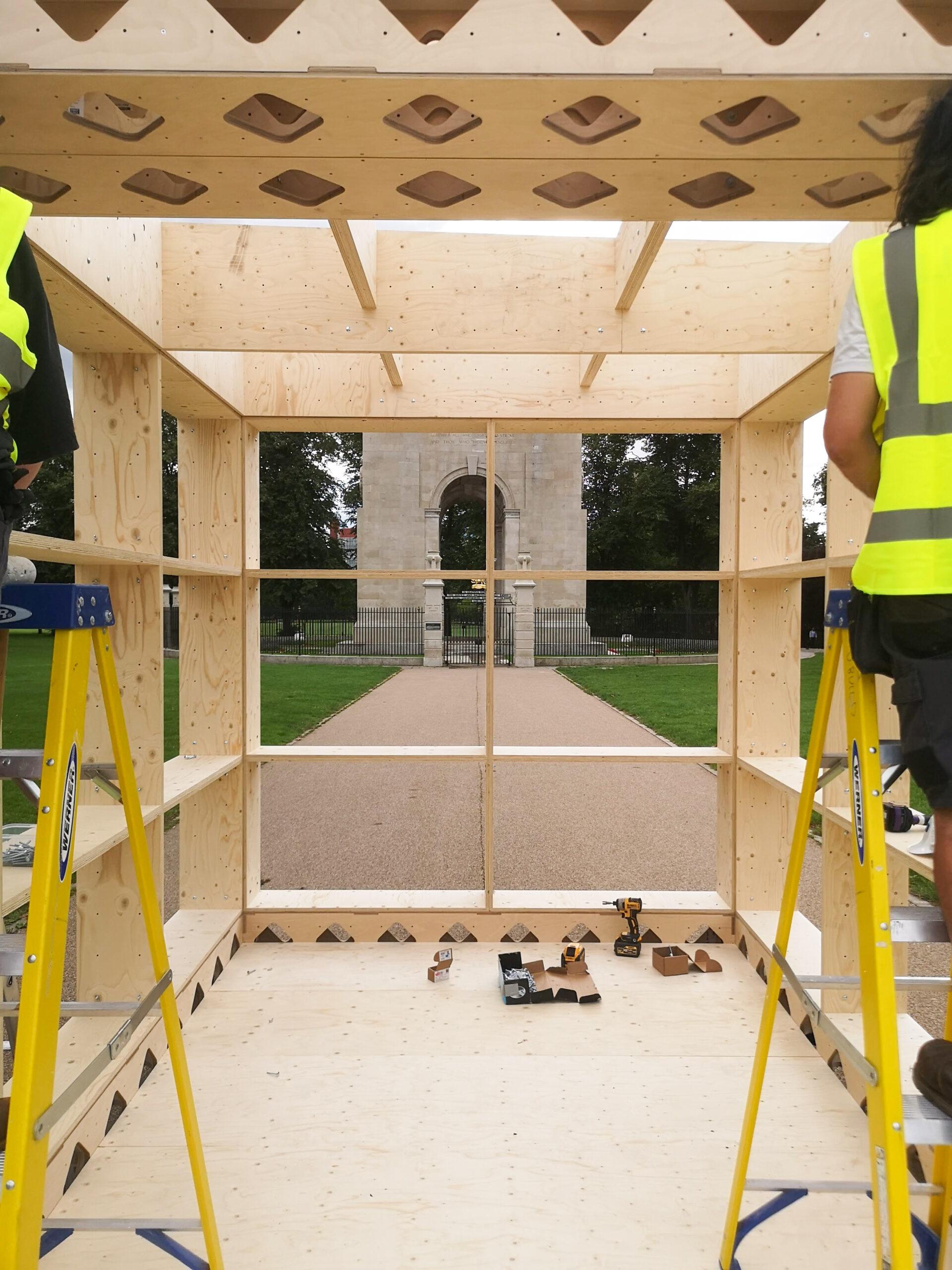 View from inside a partially constructed modular wooden structure, with two people in yellow safety vests standing on ladders at each side. An arch monument and pathway are visible through the window at the far end.