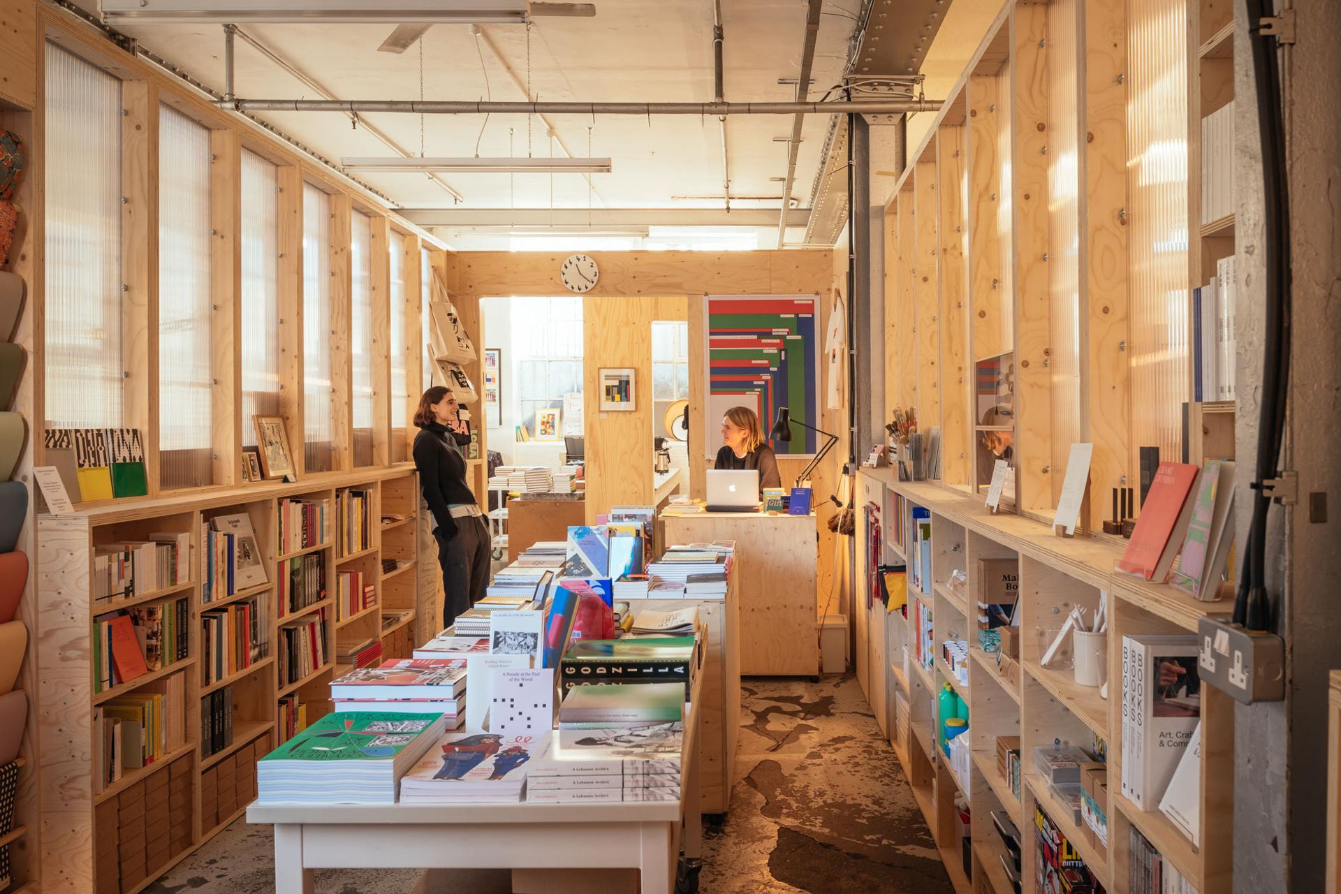 A cosy, well-lit bookshop with sustainable birch wooden shelves full of books and magazines, tables displaying colourful books, and two people talking at the counter in the background under a wall clock.