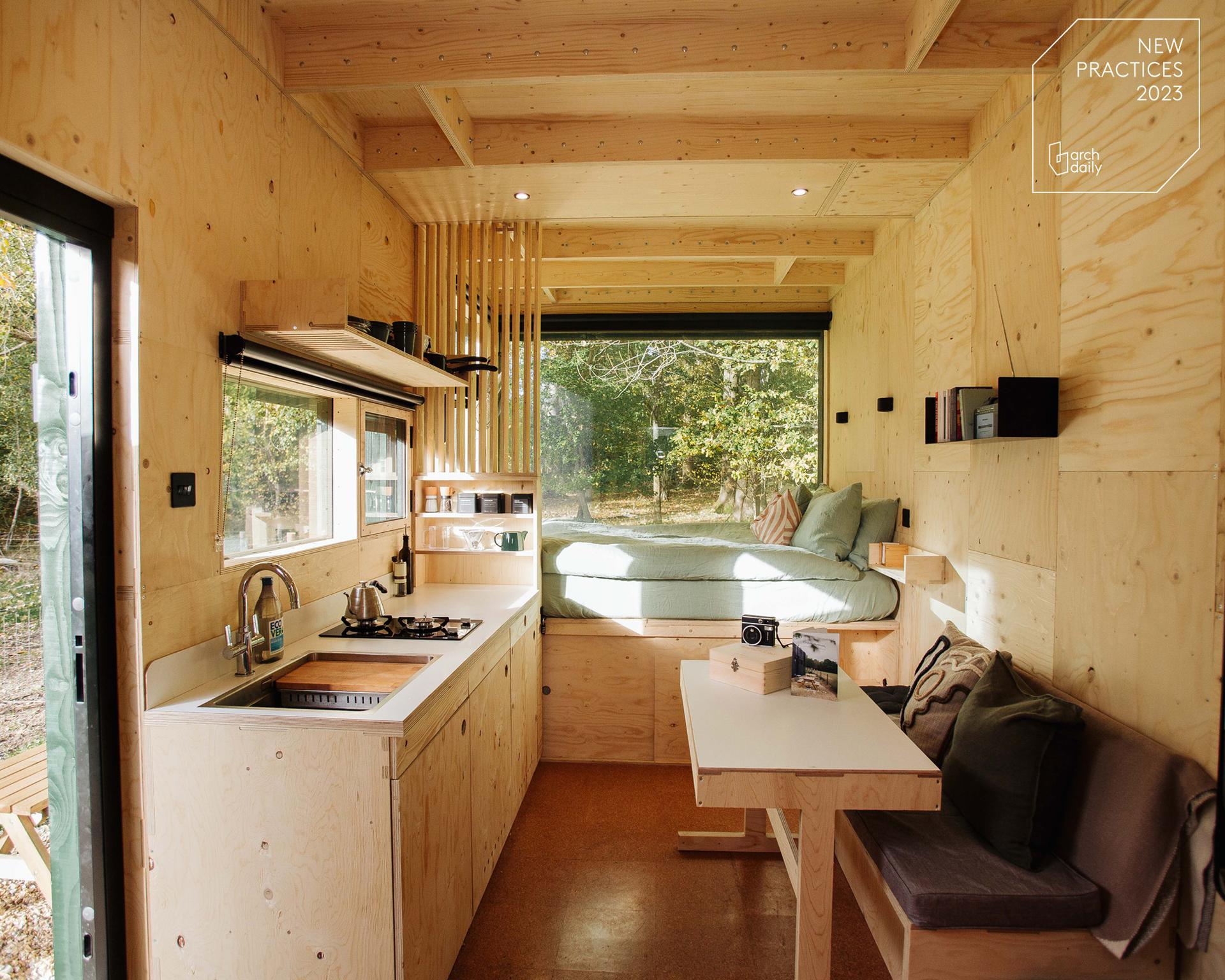 A bright, modern tiny house interior with light spruce wood walls, a kitchenette, a dining table, a modular built-in bed by a large window, and shelves. Sunlight streams in, and lush greenery can be seen outside.