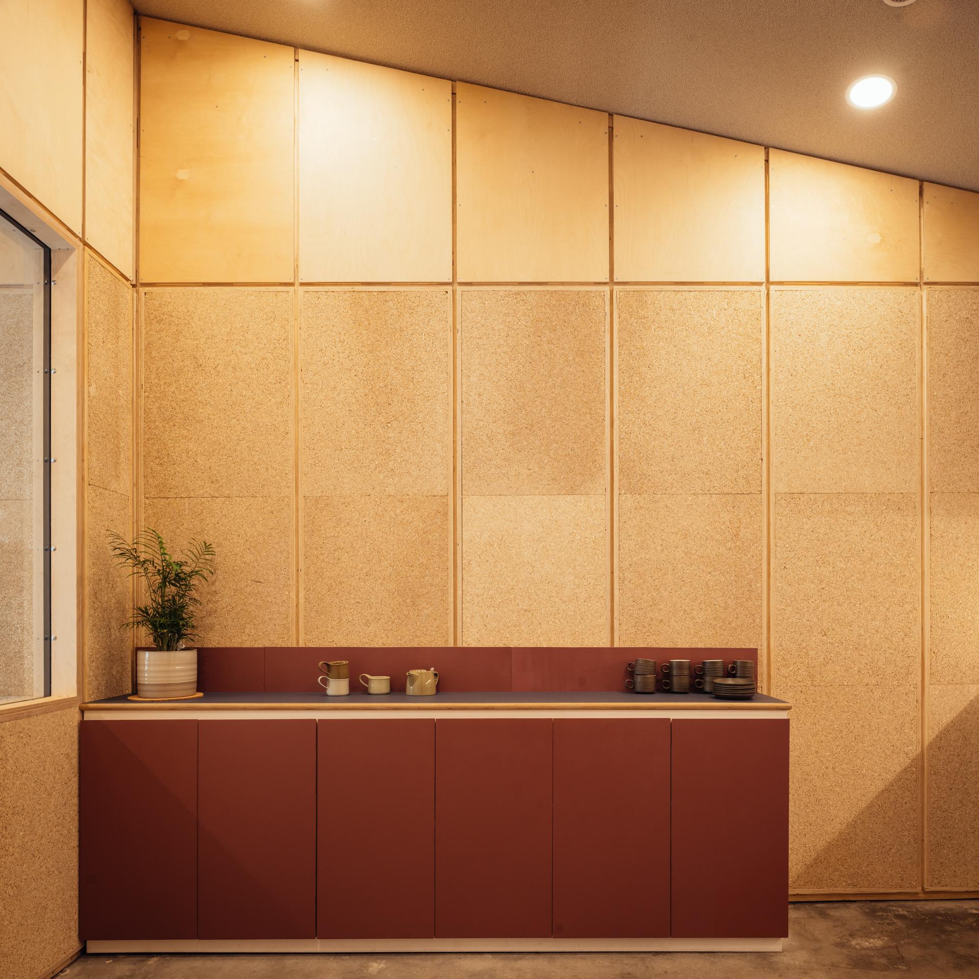 Minimalist kitchenette at Vyner Studios with red lower cupboards, beige cork walls, a plant in a striped pot, cups, and teapots on the worktop. Ceiling slopes upward at an angle. Warm indoor lighting.
