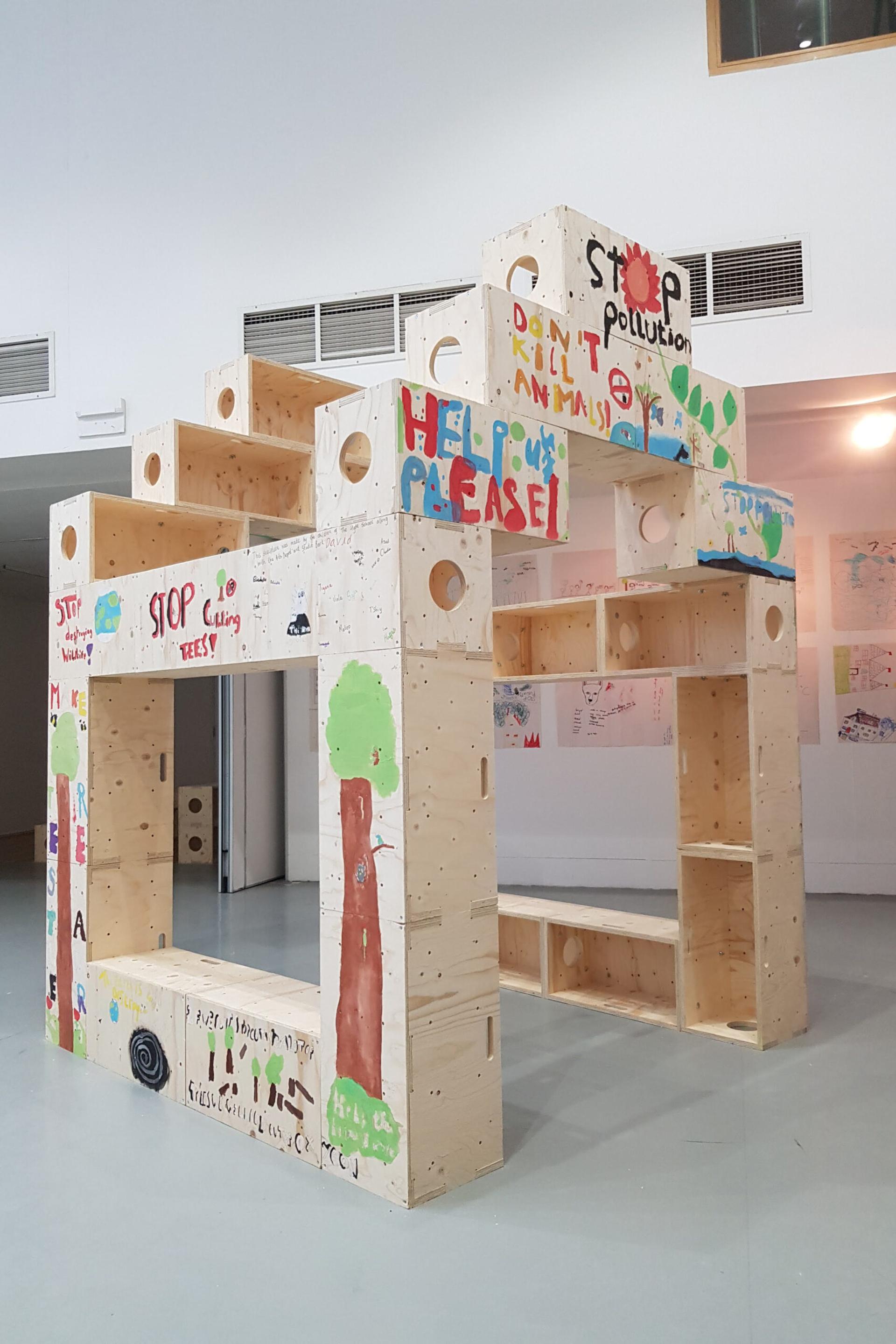 A large wooden structure indoors, painted with colourful messages and drawings about protecting the environment and promoting sustainability, including phrases like Stop pollution and Help please. Children’s art adorns the walls in the background.