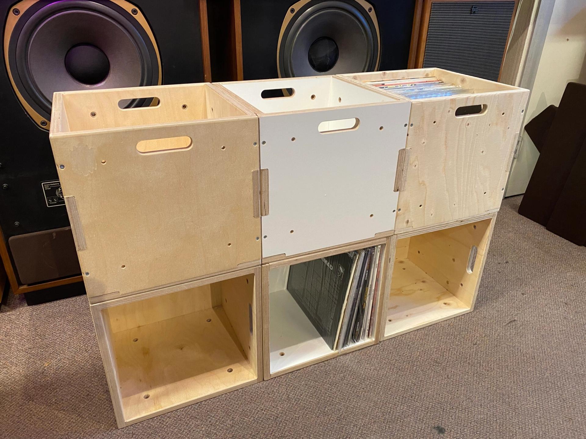 Three wooden storage crates, made for reuse, are stacked as a shelf unit holding vinyl records. Some feature white panels. Large speakers sit in the background on a carpeted floor, embracing the spirit of a circular economy.