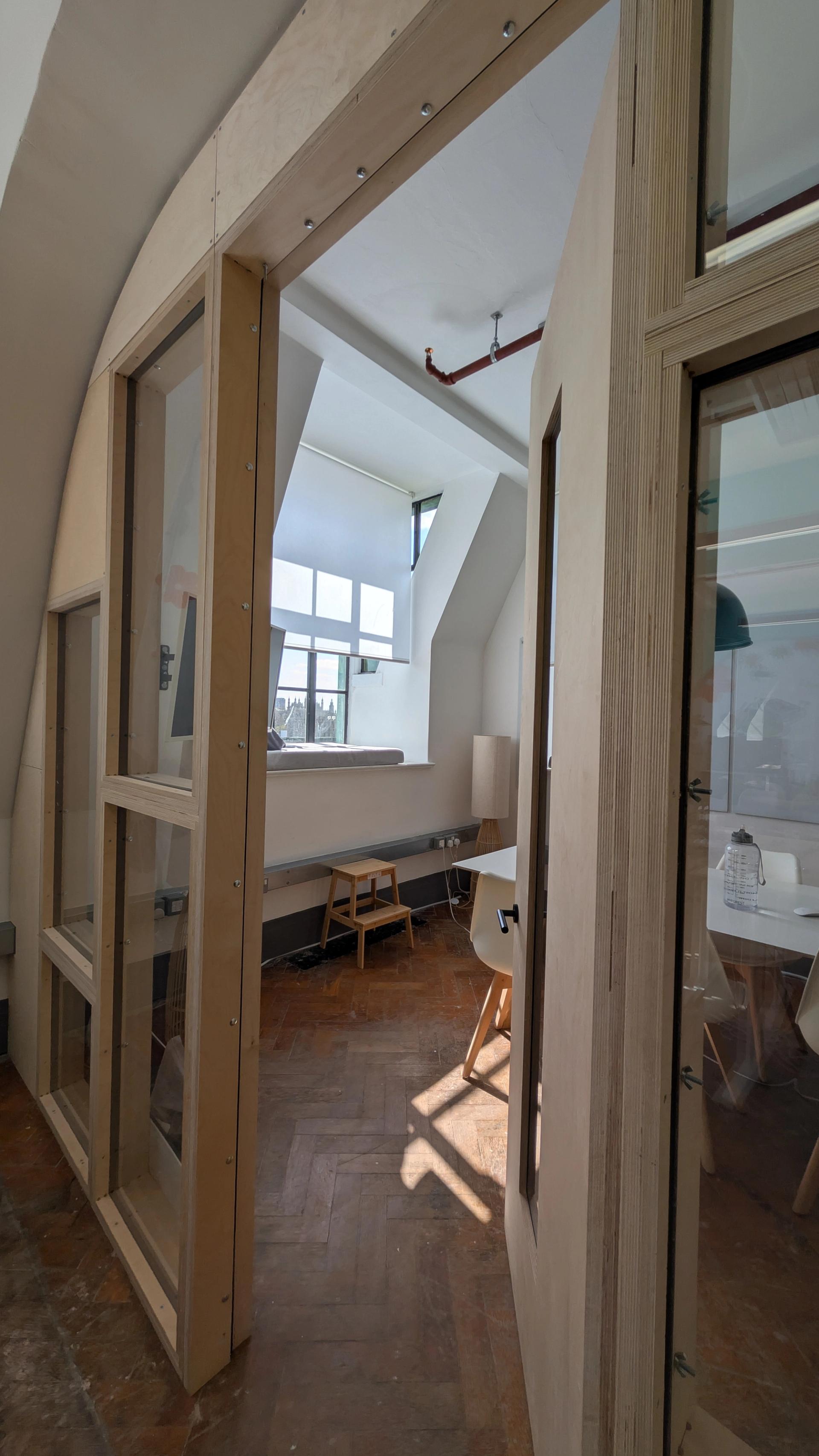A modern office space with large windows, wooden floors, and white walls showcases sustainable timber construction. Minimal furniture, including a plywood desk and chairs, complements the bright and airy atmosphere flooded with sunlight.