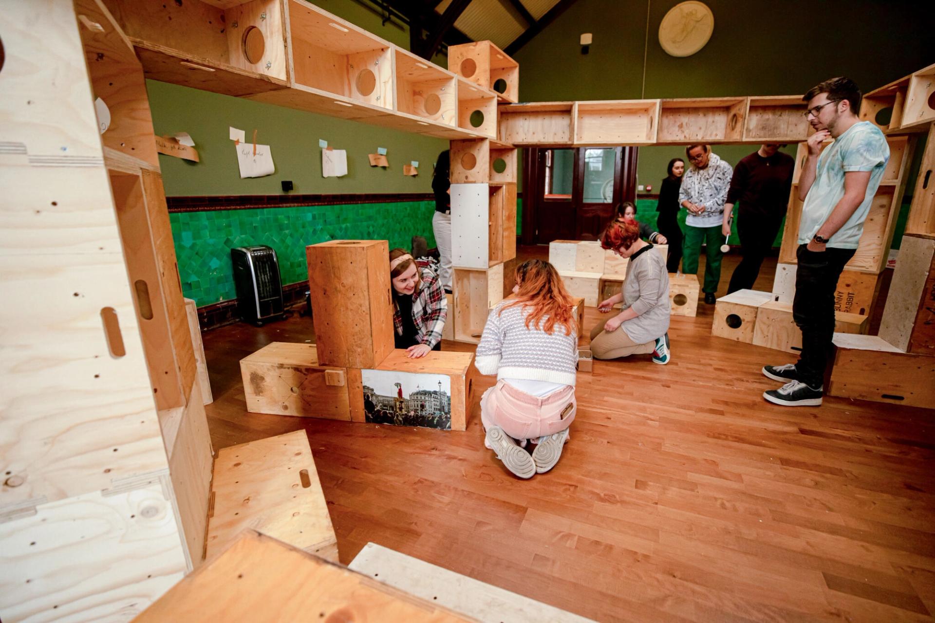 Several people interact with a large birch plywood block structure in a spacious room with wooden floors and green walls. Some are kneeling or crouching, while others stand and observe. The sustainable design encourages hands-on engagement.