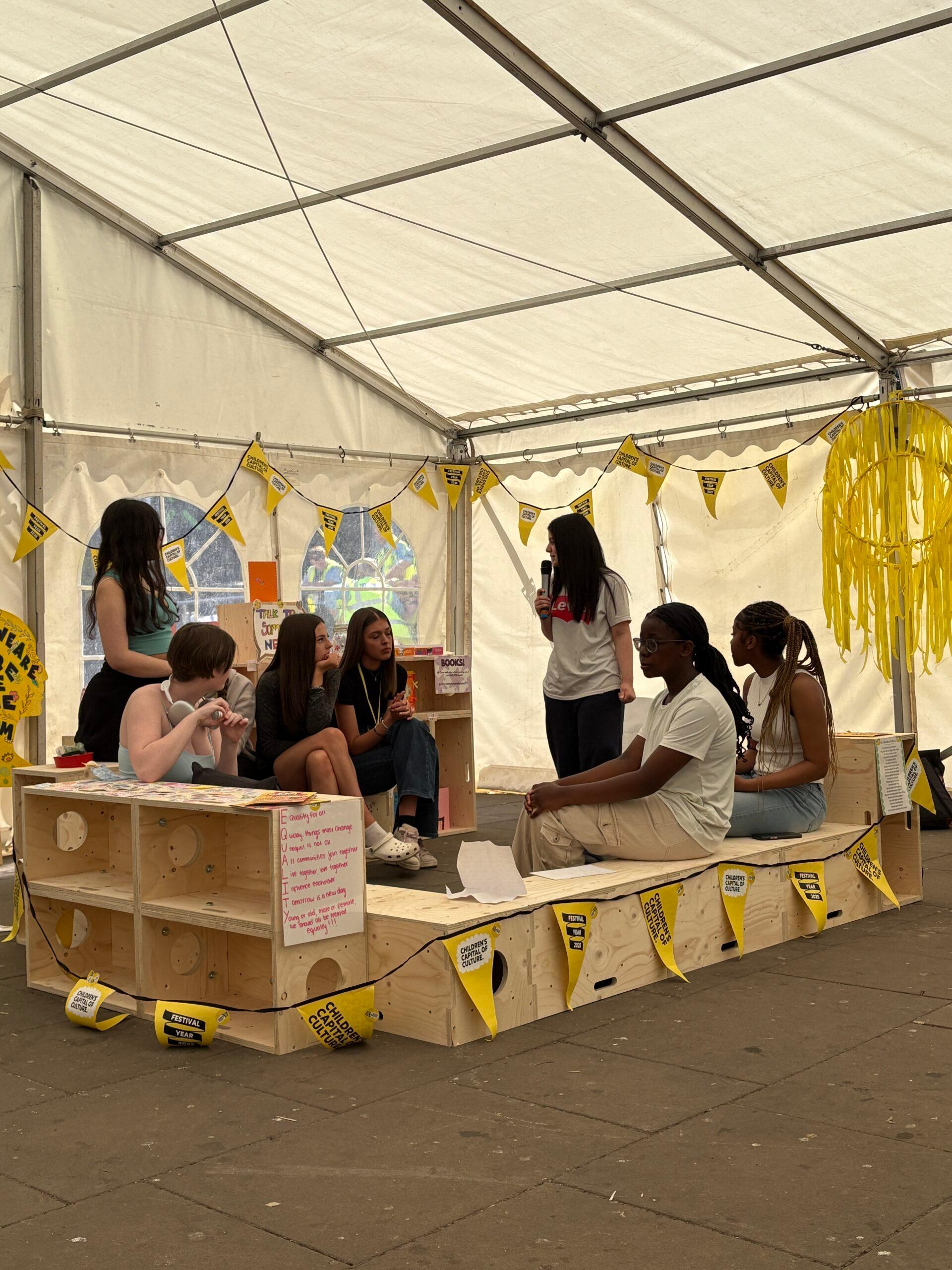 Six young people sit and talk on birch benches inside a white marquee decorated with yellow banners and streamers; one person stands and speaks, holding a microphone.