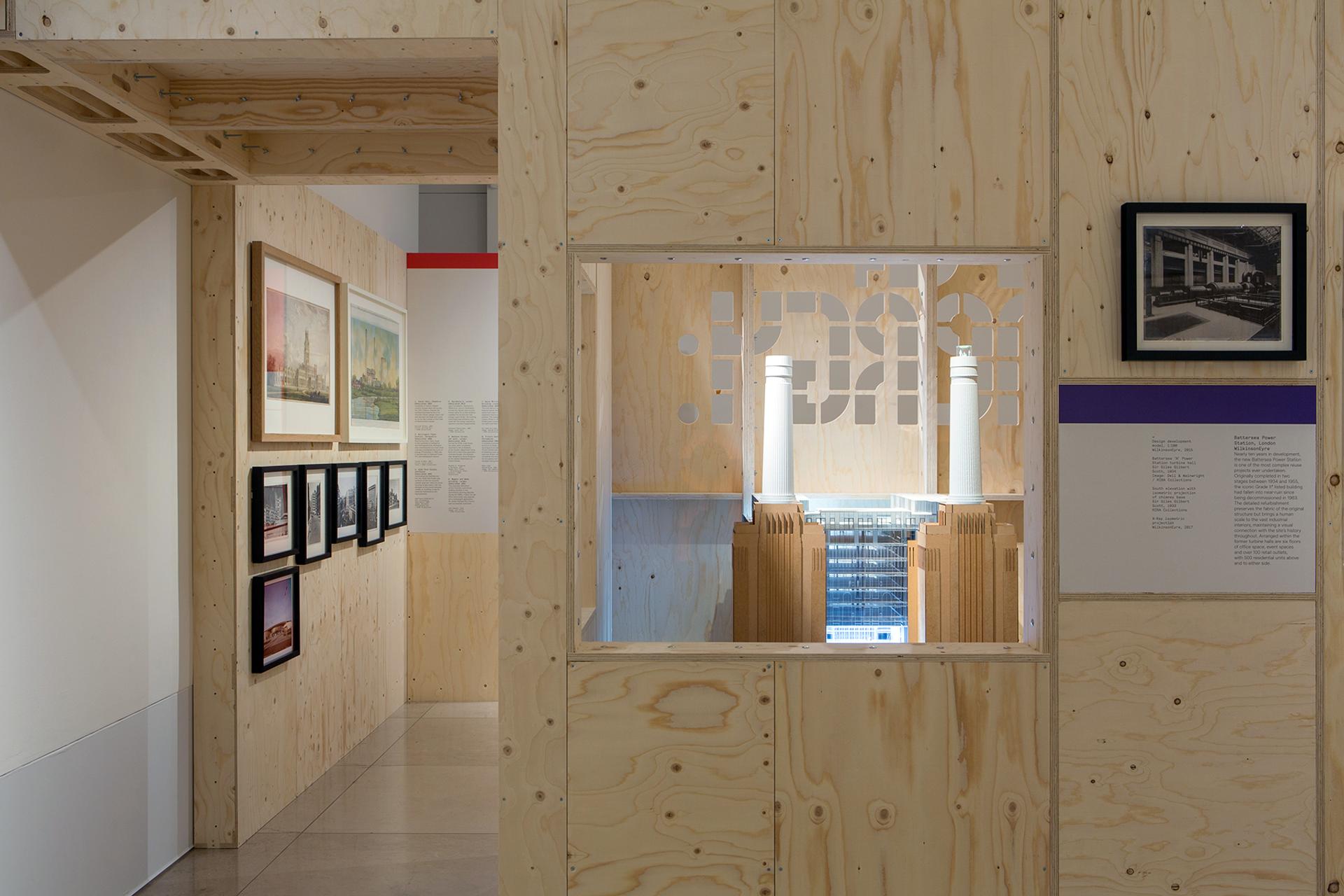 A room with light birch plywood walls displays framed photographs and artwork. A square window reveals another space with architectural models and vertical white pipes. Text panels are mounted beside the window and on the walls.