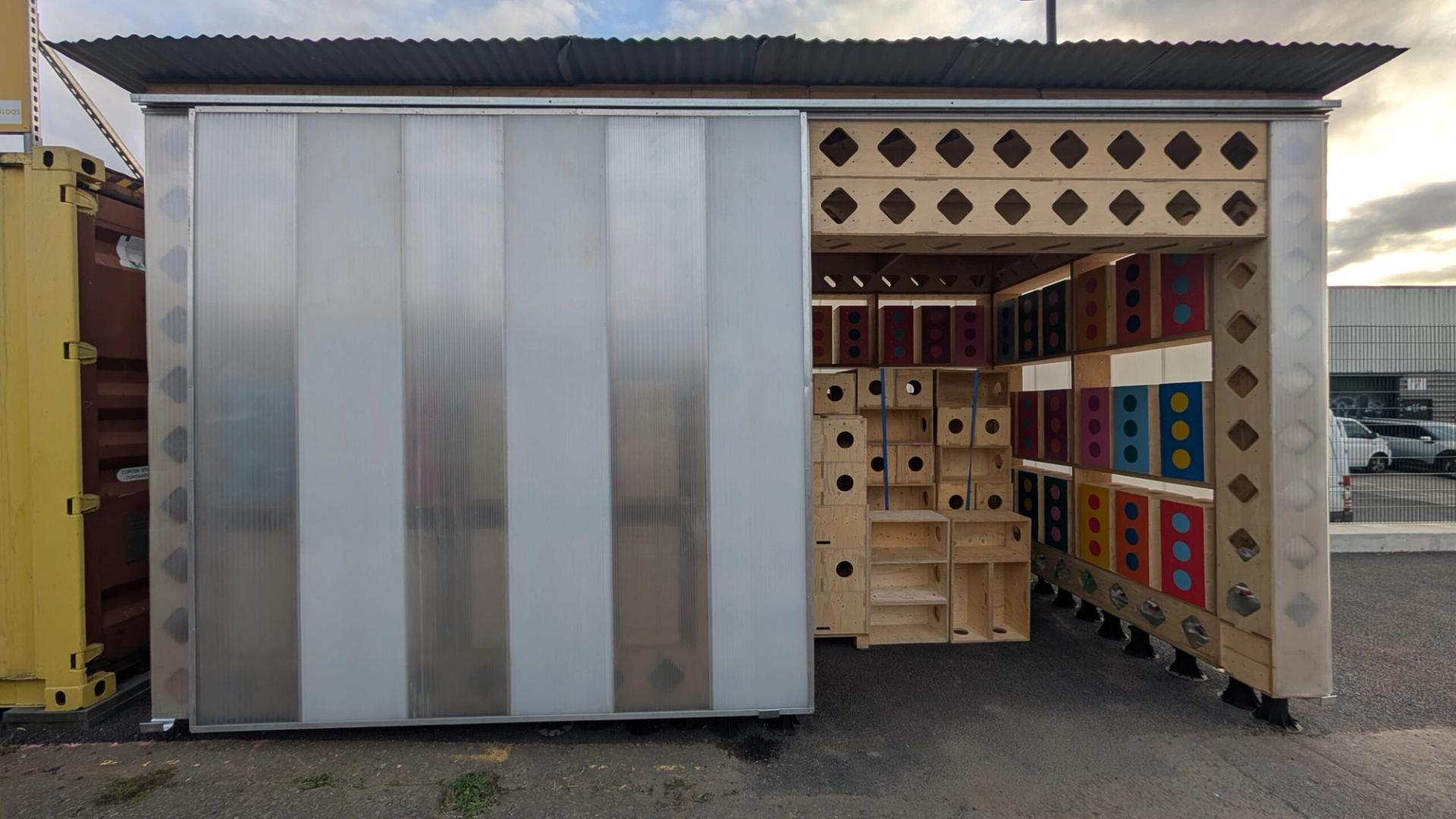 A small U-Build wooden structure with patterned cut-outs and open sides, featuring stacked wooden boxes with colourful circular holes inside. The setting appears to be an industrial outdoor area.