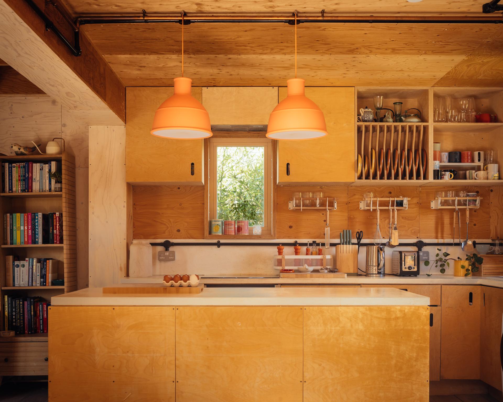 A kitchen with plywood cupboards and a central island within a box house extension, featuring two bright orange pendant lights above the worktop and a bookcase on the left.