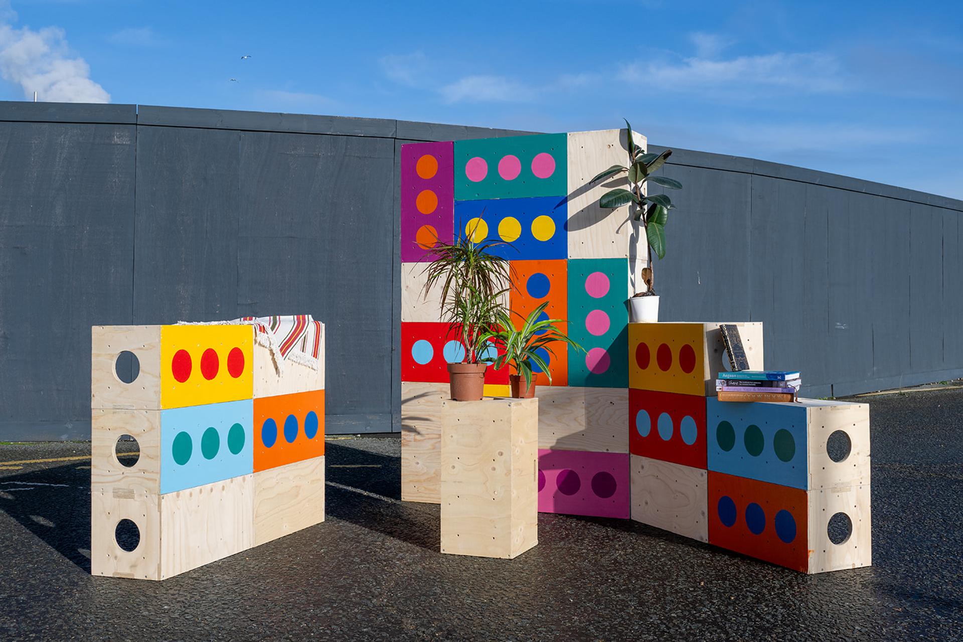 Colourful wooden furniture and panels with painted circles are arranged outdoors on asphalt, capturing the charm of a box library among potted plants, books, and a grey fence in the background under a blue sky.