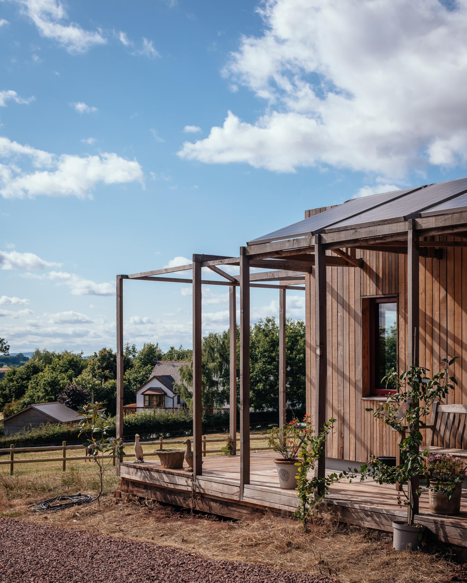 A modern wooden house featuring birch timber construction, a partially covered veranda with potted plants and a chair, set in a rural area surrounded by green trees, grass, and other houses under a blue sky with scattered clouds.