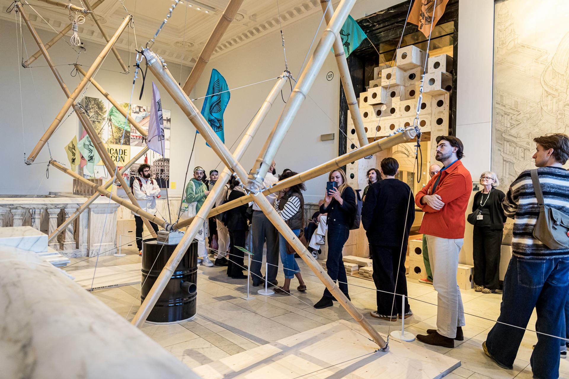 A group of people stands around a large plywood geometric installation in a museum gallery, observing and discussing the exhibit. The room has high ceilings, artwork, and architectural decorations.