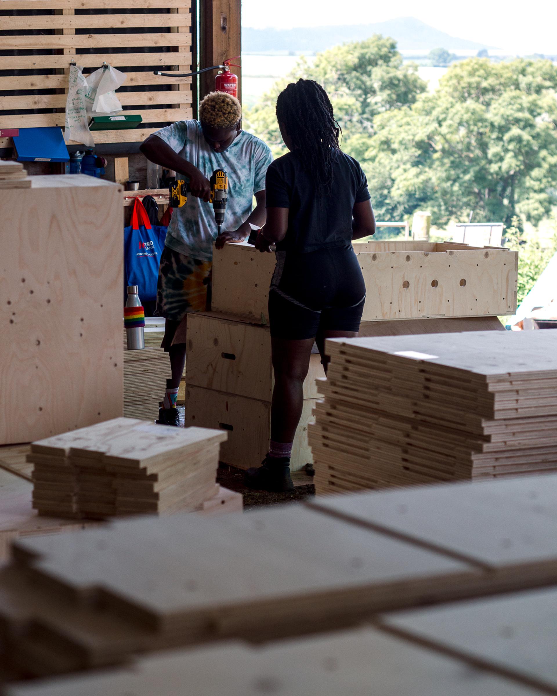Two people assemble wooden furniture in a workshop filled with stacked, sustainably sourced plywood pieces. One person uses a power drill whilst the other holds the wood steady. Sunlight streams in, and trees are visible through the open entrance.