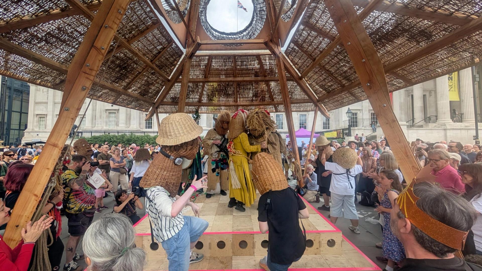 A crowd watches performers wearing large woven straw masks and hats under a wooden pavilion in an outdoor public square, as people take photos and clap—capturing the spirit of the 2025 Triumph of Art at the National Gallery.