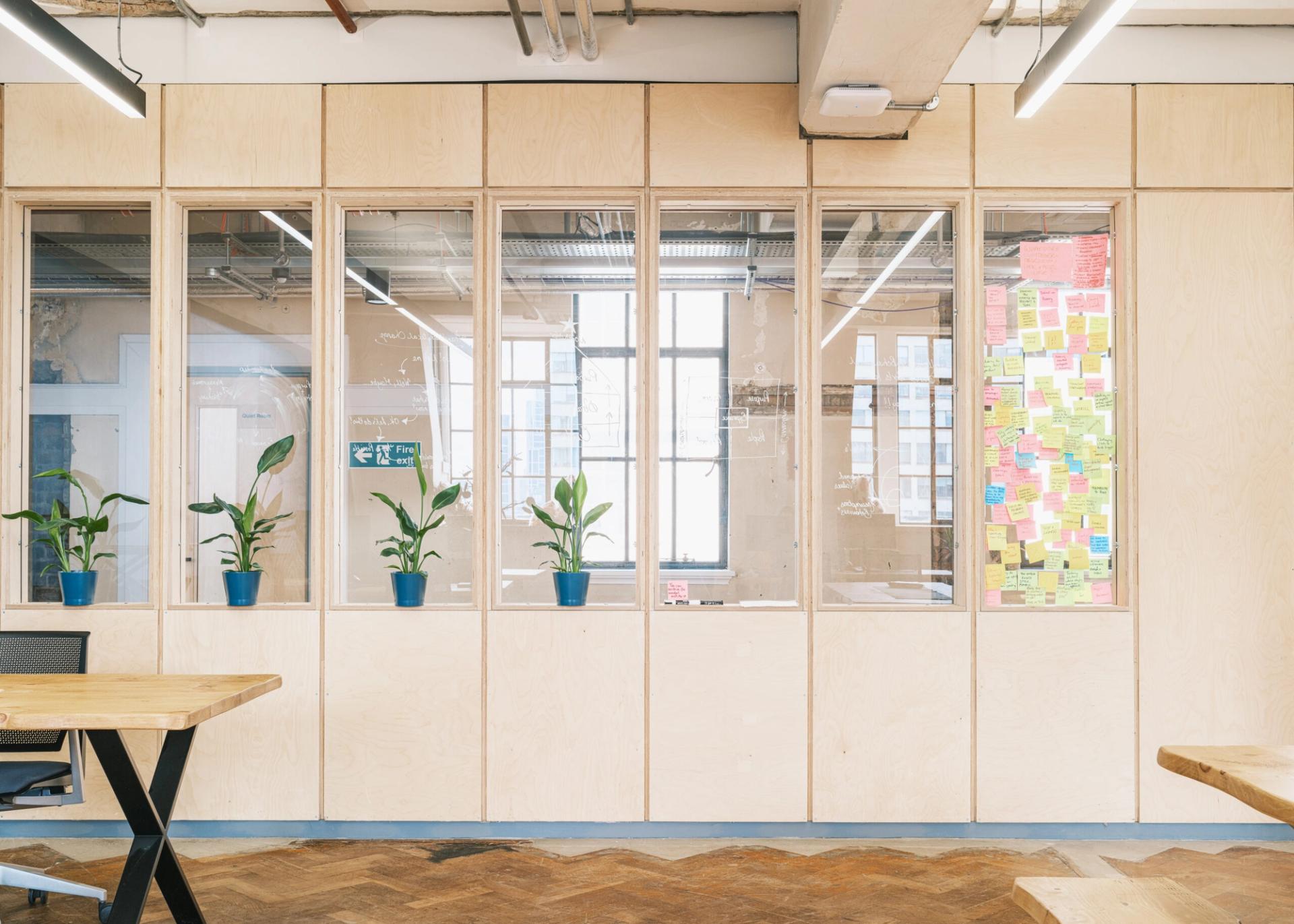 Modern office space with birch plywood walls, five potted plants on a ledge, large windows letting in natural light, and a glass panel covered in colourful sticky notes. Modular tables and chairs are partially visible in the foreground.
