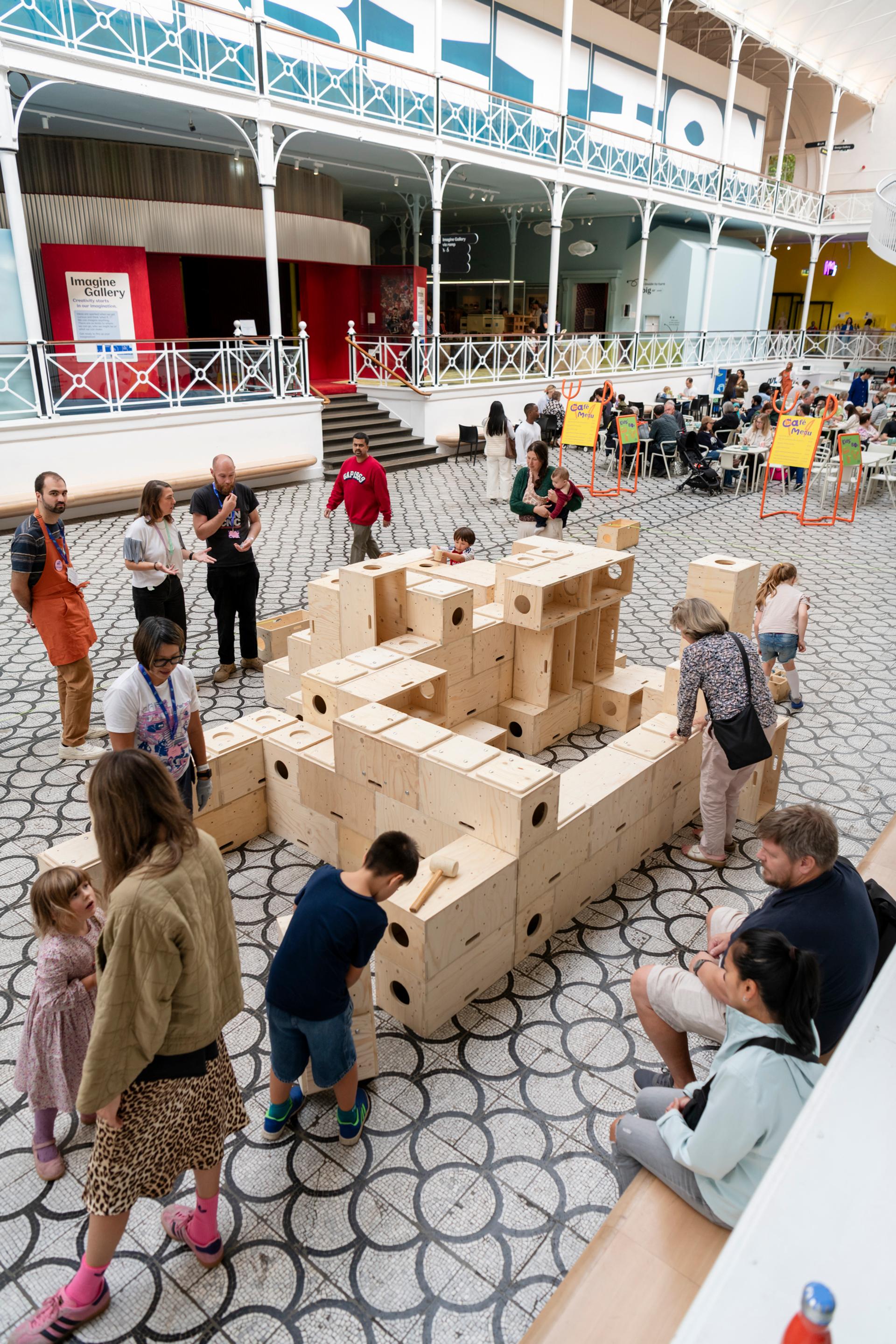 People interact with large wooden U-Build blocks stacked in various shapes inside a spacious, bright museum with patterned floors and a high ceiling. Some adults and children are building, whilst others watch or walk by.