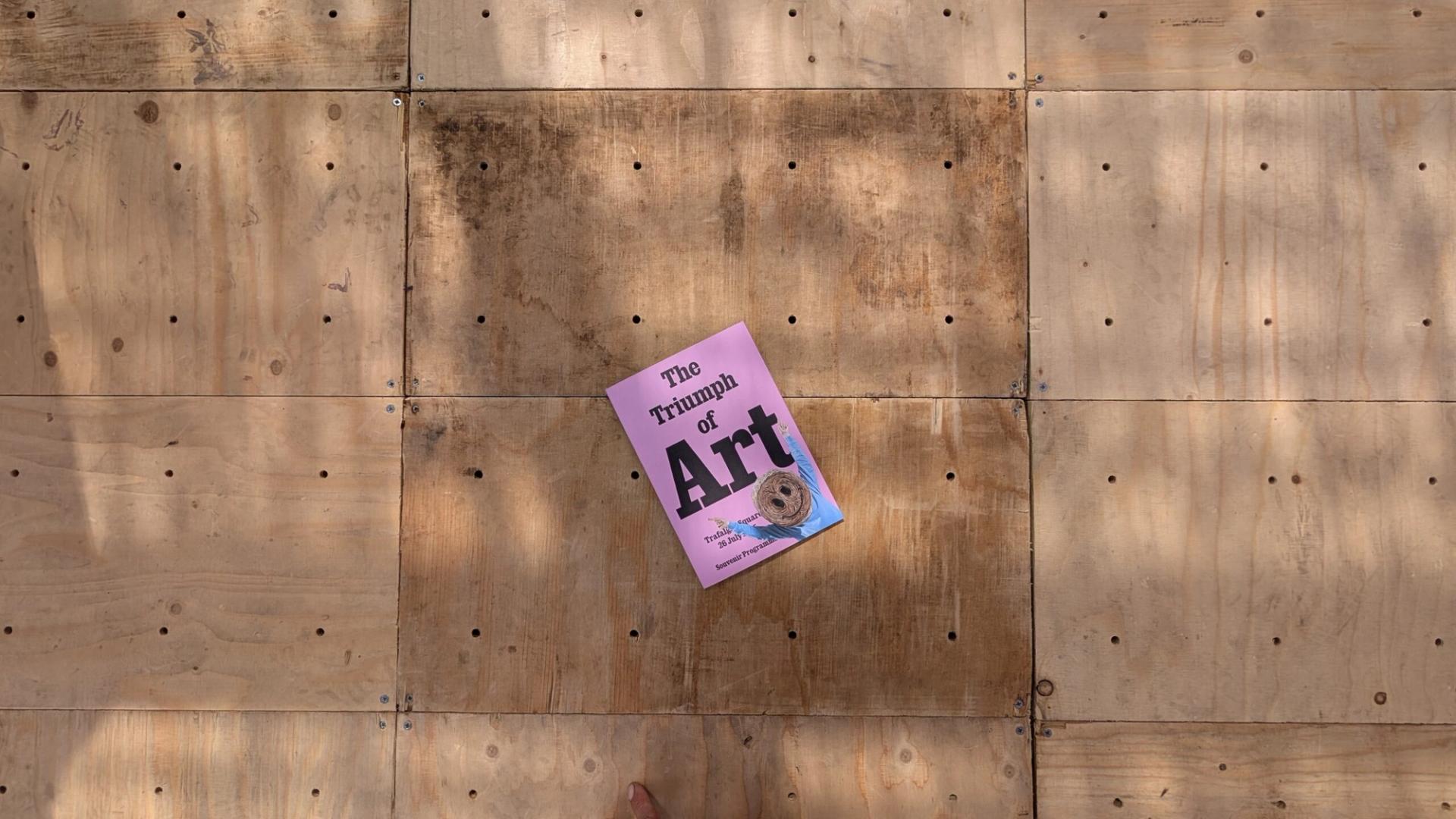 A pink book titled The Triumph of Art lies on a wooden surface made of square panels, showcasing visible screws and stains—hinting at reuse or the circular economy. A partial finger is visible at the bottom edge of the image.