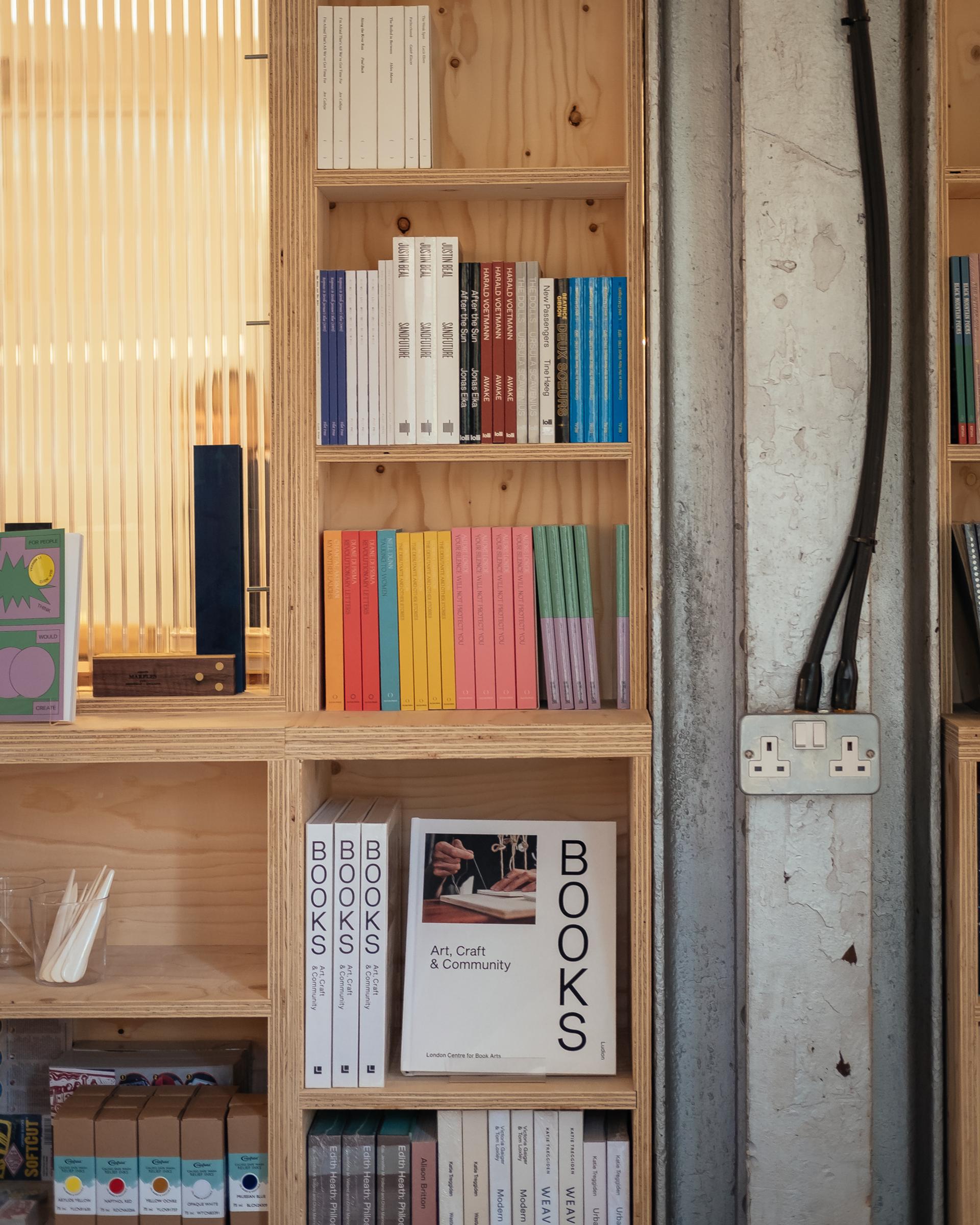A sustainable plywood bookcase displays neatly arranged books, including colourful spines and large books titled BOOKS: Art, Craft & Community. An electrical socket and exposed cables are visible on the right side of the shelf.