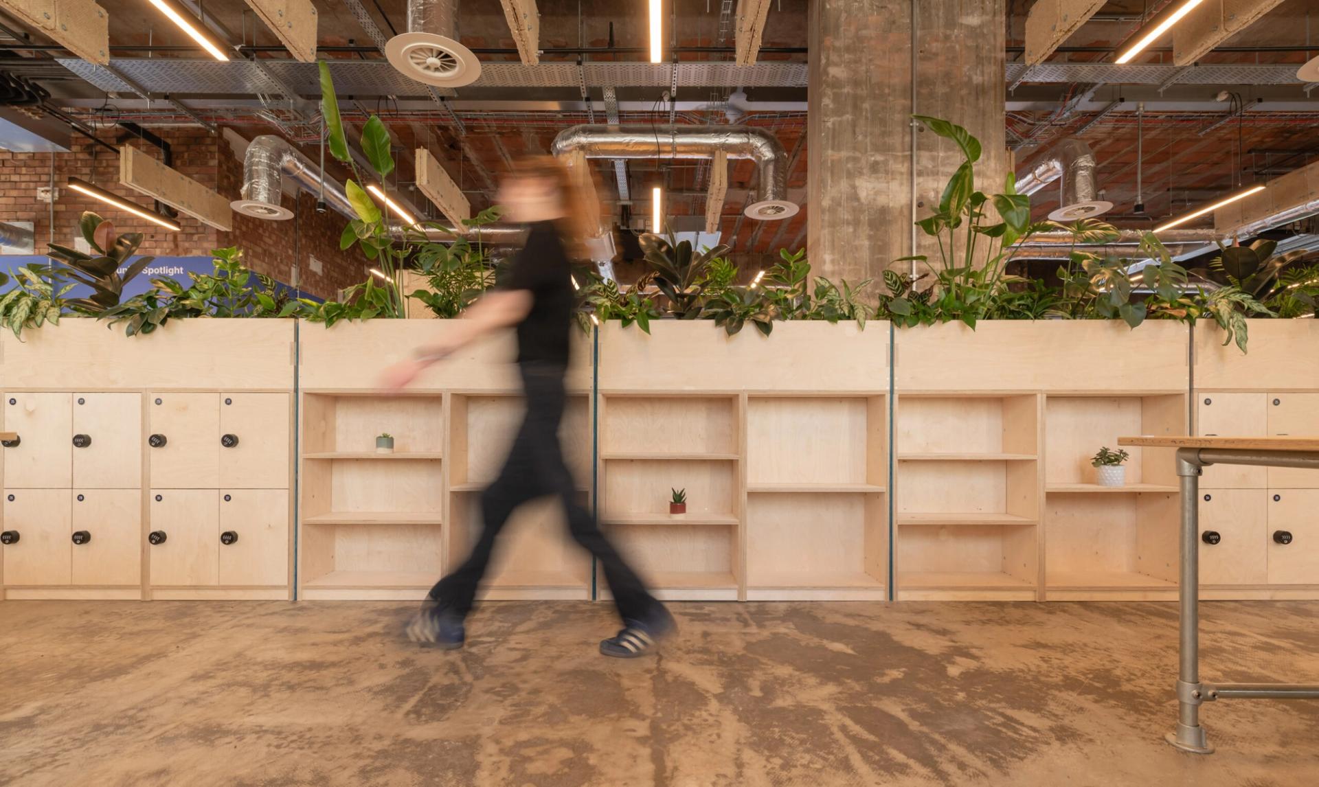 A person in motion walks past wooden shelves and lockers in the Manchester workspace of Sustainable Ventures, featuring exposed pipes, plants, and a concrete pillar—a hub for modern industrial design and sustainable business innovation.