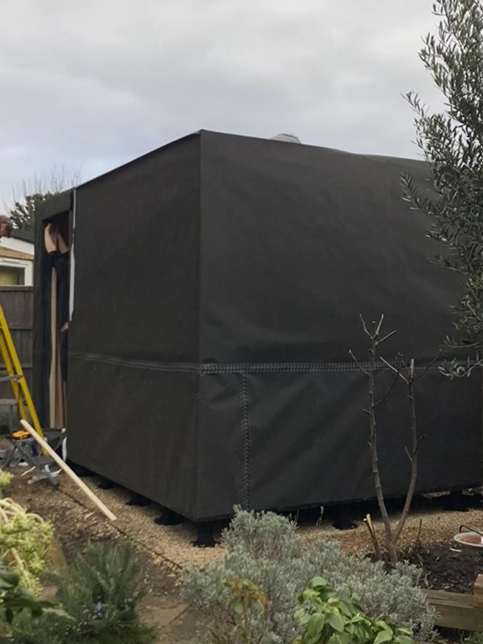 A large, black, modular box-shaped structure stands in a garden area with plants and a yellow ladder nearby under a cloudy sky.