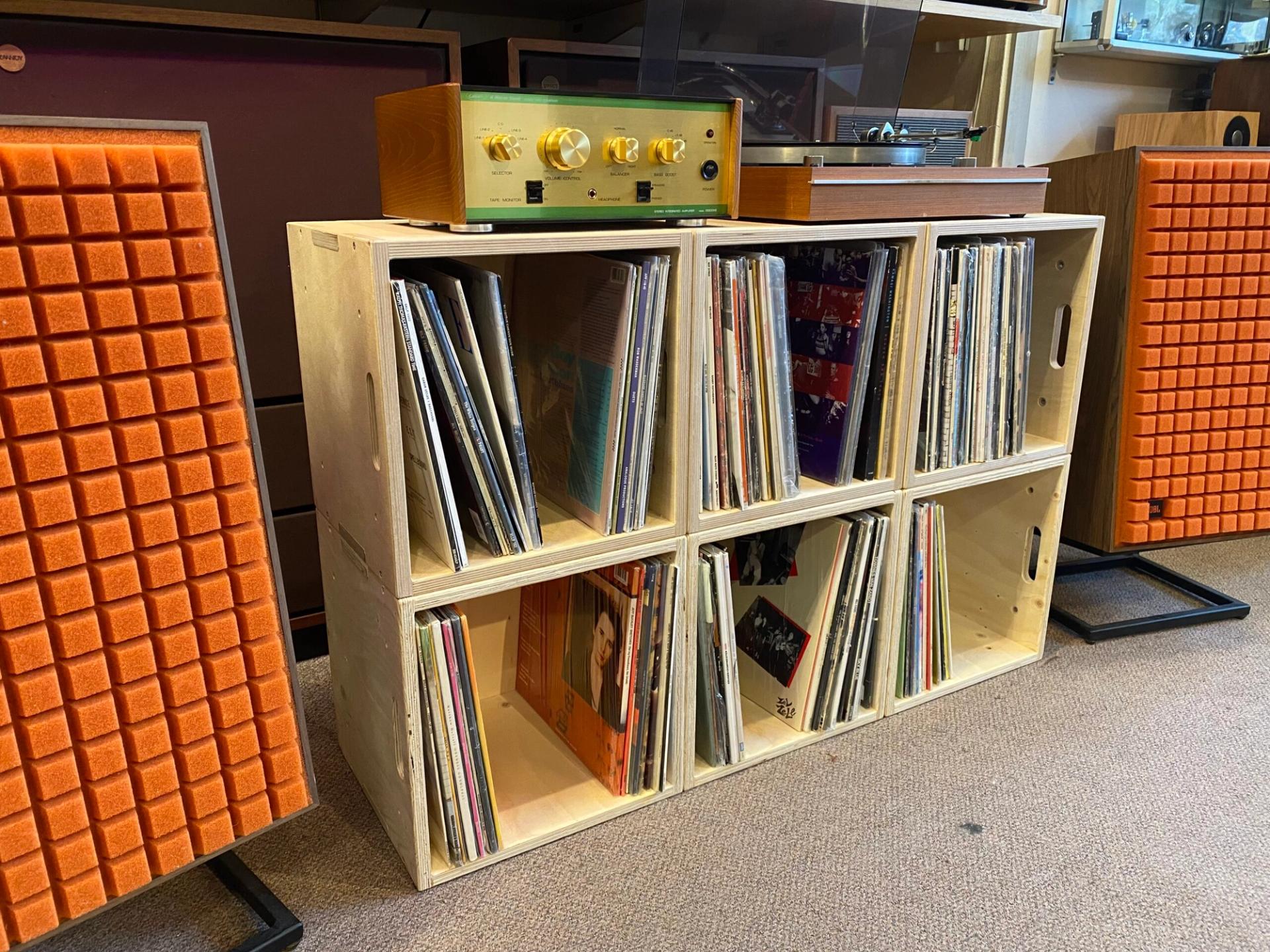 A wooden shelving unit, crafted from sustainably sourced materials, holds vinyl records and a vintage stereo amplifier with a turntable on top. Orange speakers with textured fronts stand on either side on a carpeted floor.