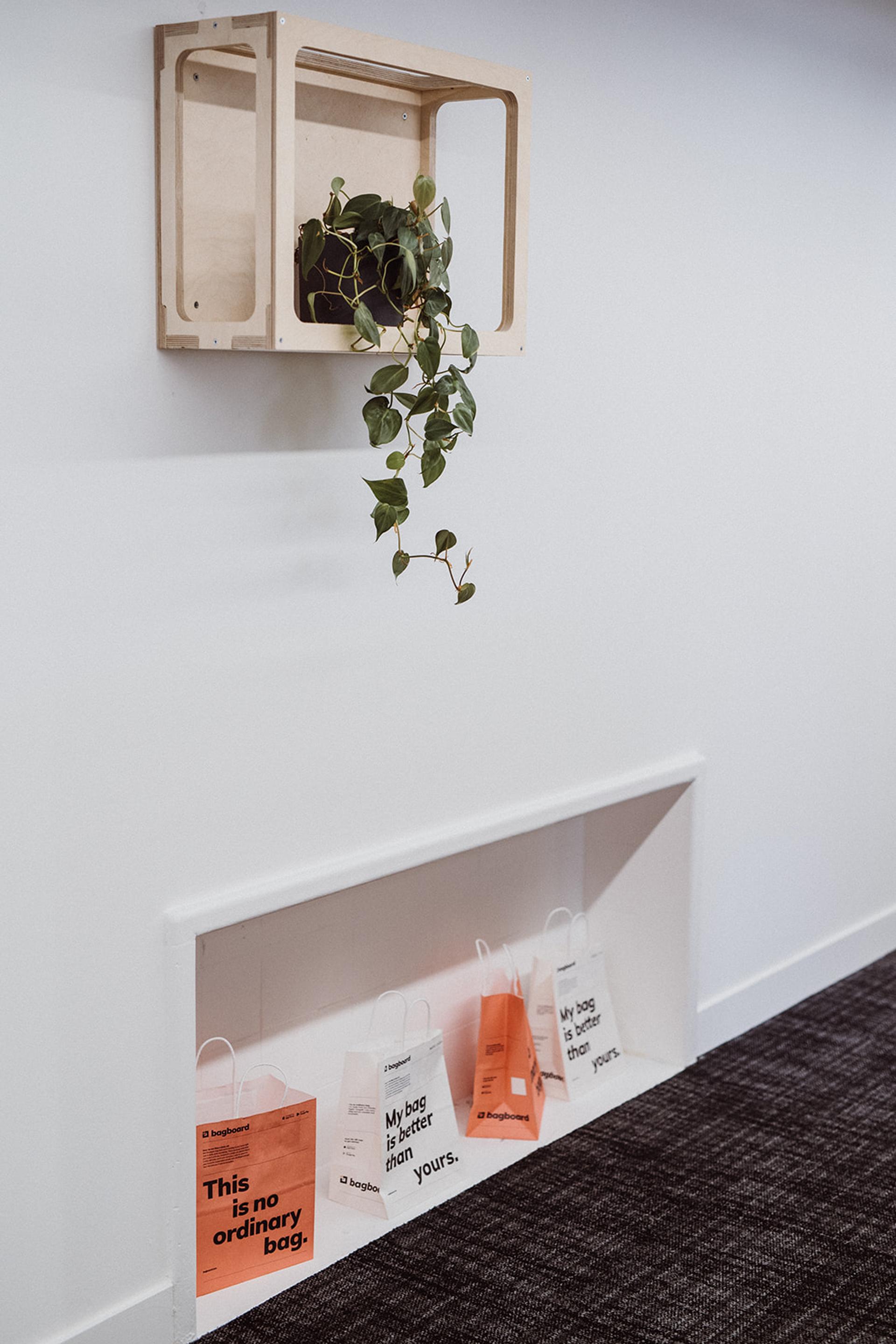 A plant on a wooden birch wall-mounted shelf hangs above a recessed alcove holding four colourful shopping bags with printed slogans on a white wall in a modern space with dark carpet.