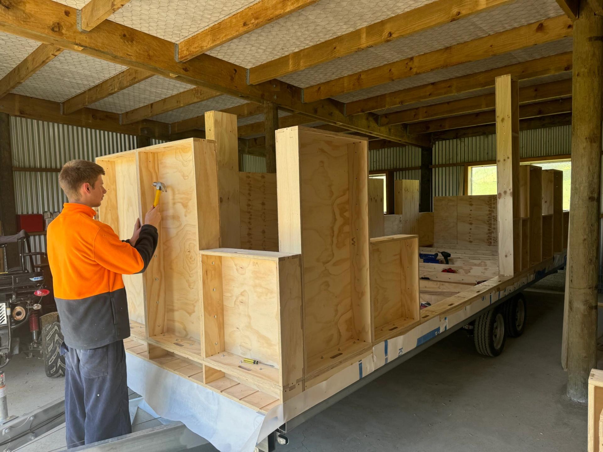 A person wearing an orange and black jacket hammers nails into a birch plywood structure being built on a trailer inside a large shed with wooden beams and corrugated metal walls.