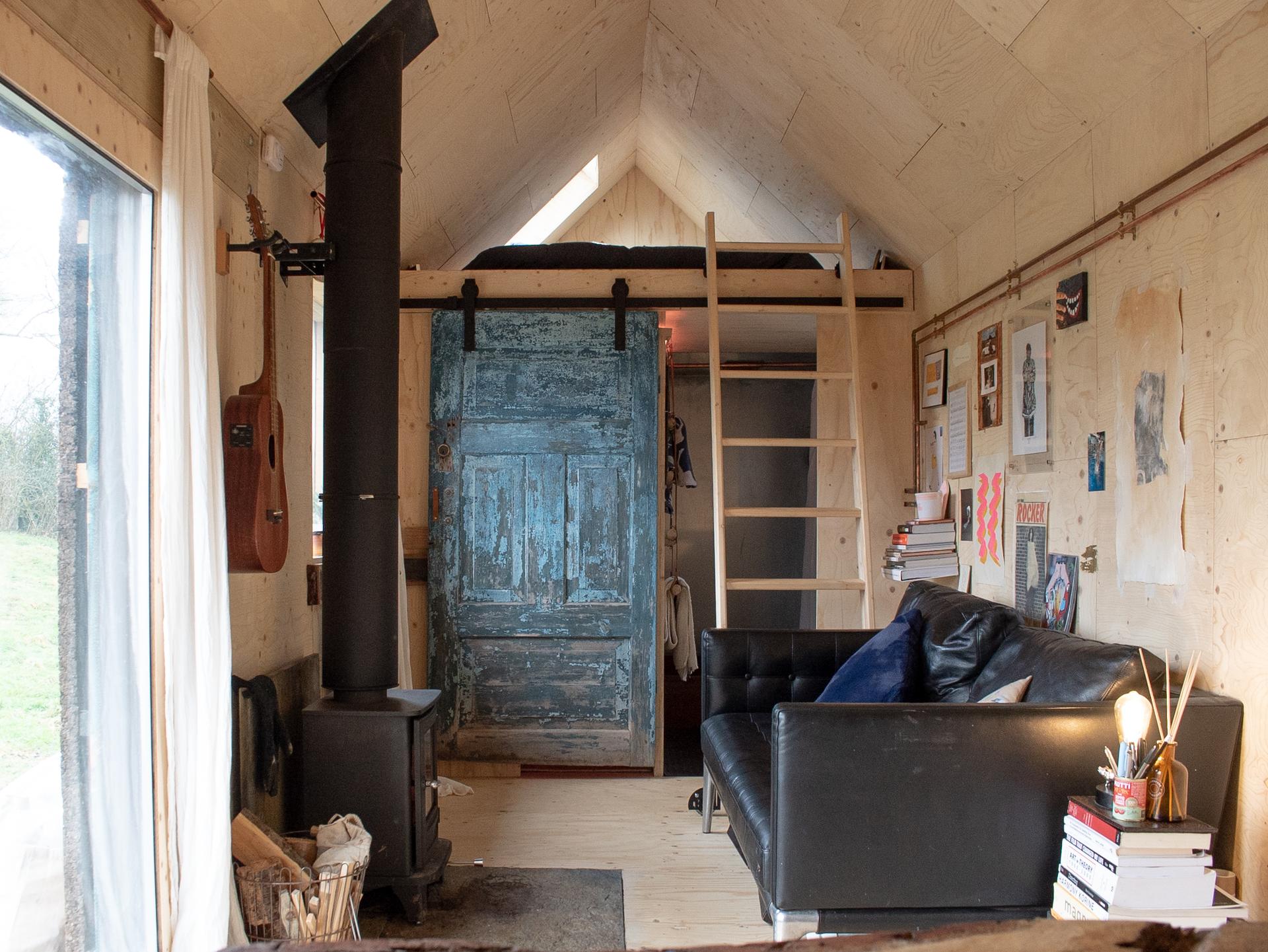 A cosy, rustic tiny house interior features a black wood-burning stove, a vintage blue door, a black leather sofa, and walls of light spruce plywood. A lofted bed with ladder, wall-mounted guitar, bookshelves and art complete this timber construction retreat.