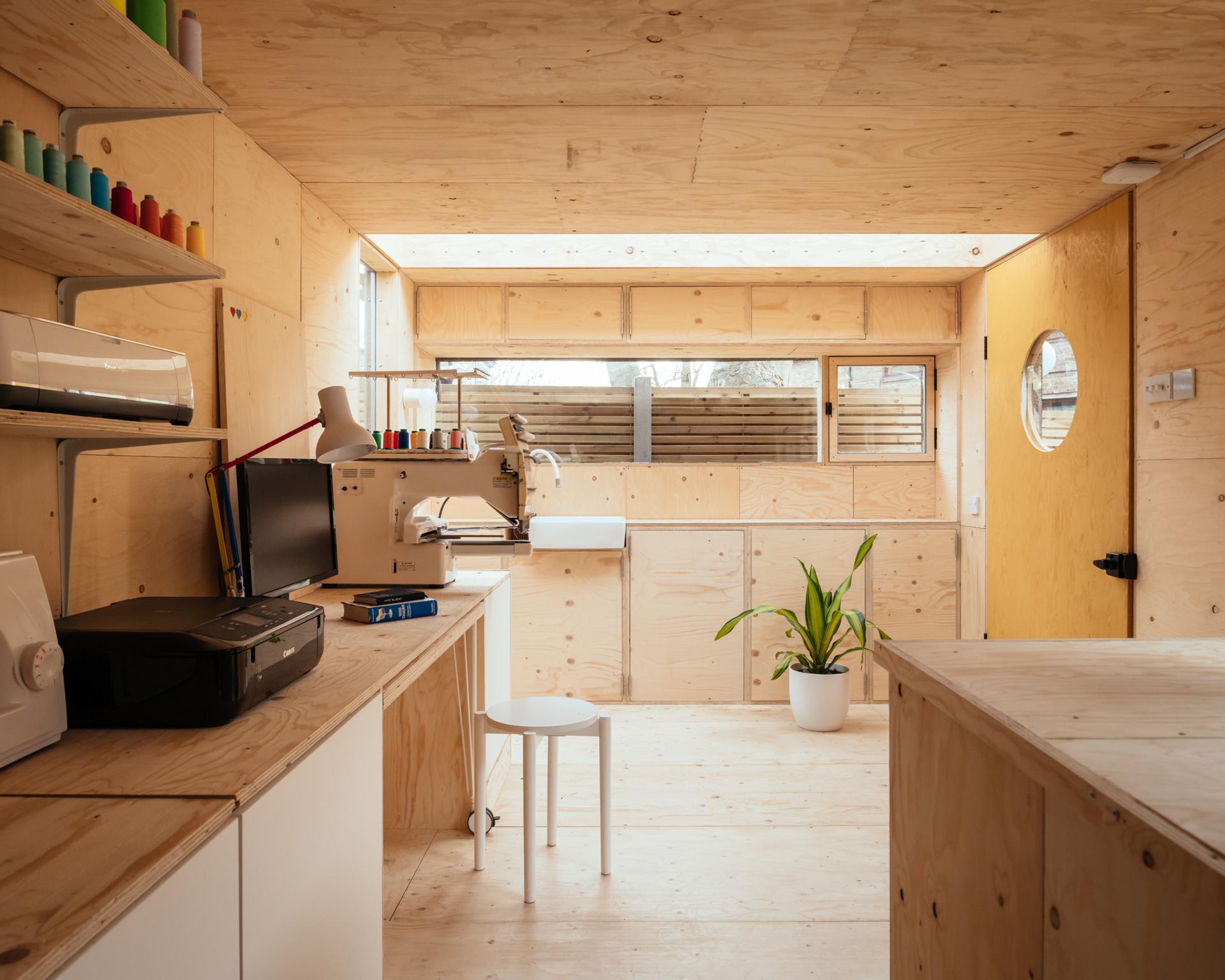 A bright, minimalist craft room with spruce wooden walls and furniture, a desk with a computer and sewing machine, modular shelves with colourful threads, a printer, a small white stool, and a potted plant near a round window.