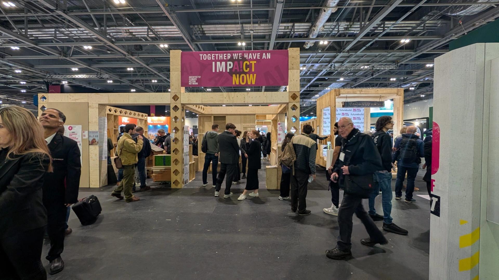 A busy indoor event with people walking and gathering around sustainable spruce and plywood stands. A large sign overhead reads TOGETHER WE HAVE AN IMPACT NOW in bold letters. The hall has high ceilings and industrial lighting.
