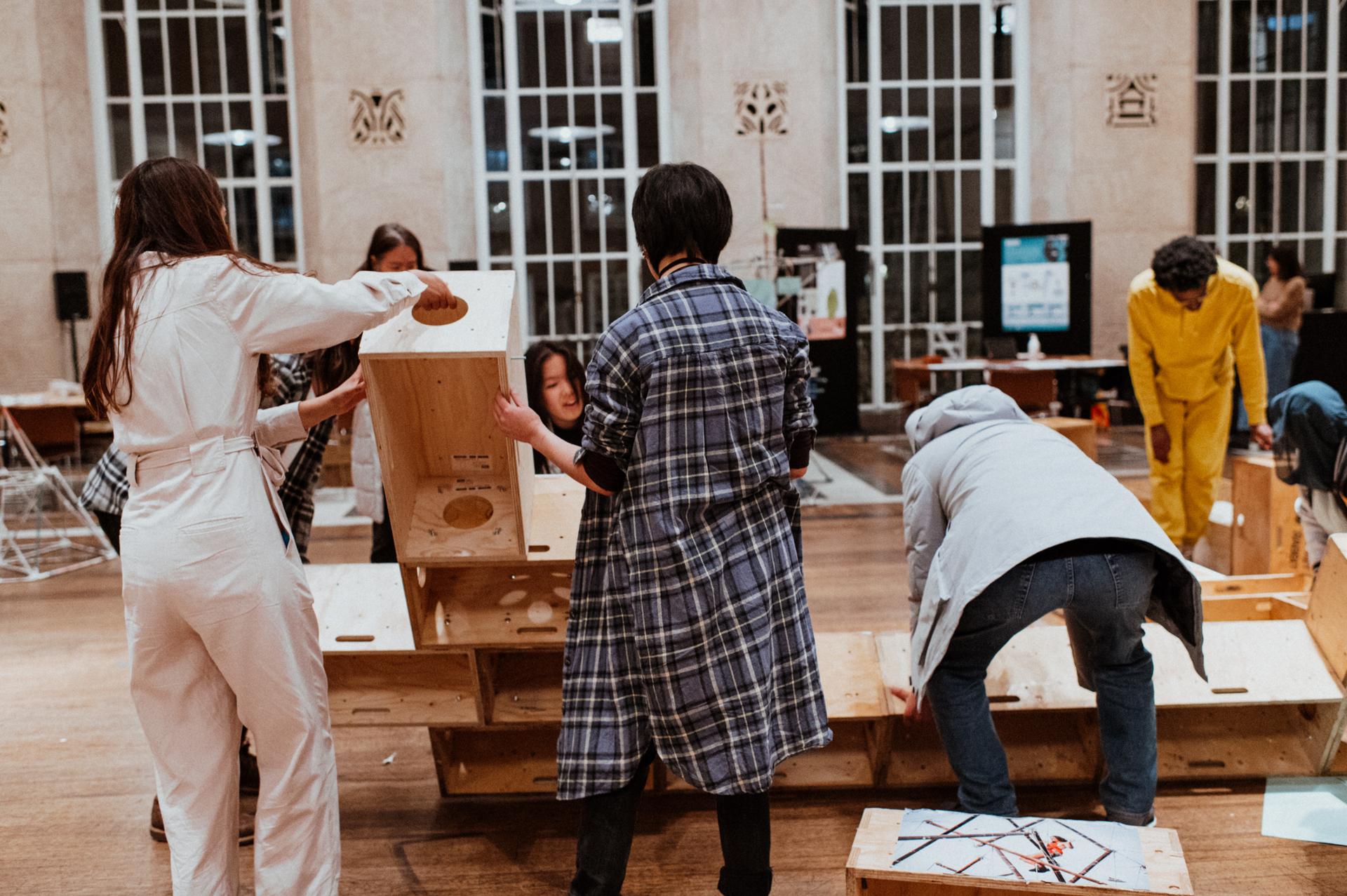 A group of people assemble modular wooden crates, some crafted from spruce and birch, in a spacious room with tall windows and wooden floors. Some lift crates whilst others bend over, working together on the project.