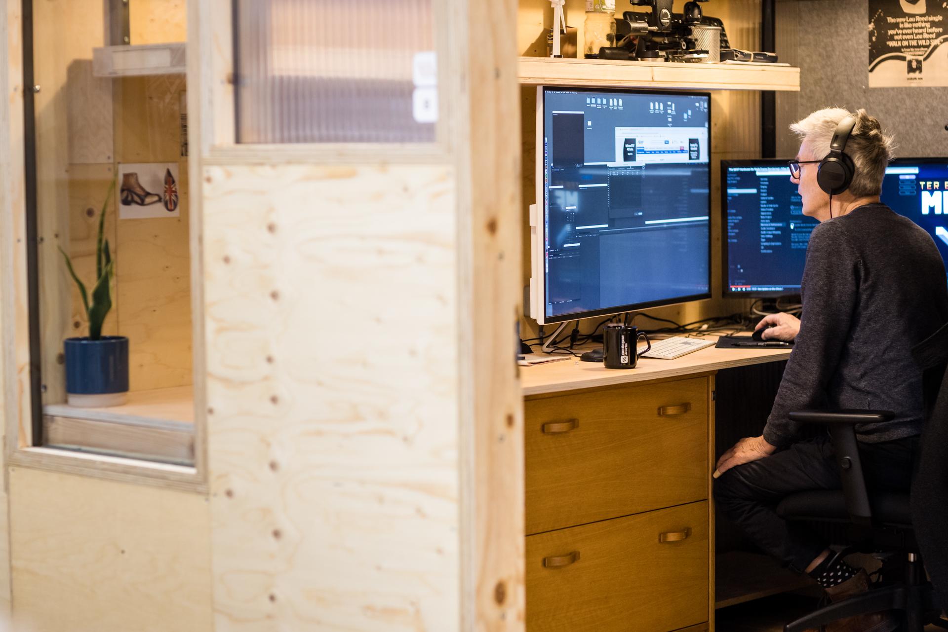 A person with headphones sits at a desk, editing video on a large monitor in a home office with sustainable birch walls. A coffee mug, a plant, and modular computer equipment are also visible on the desk.