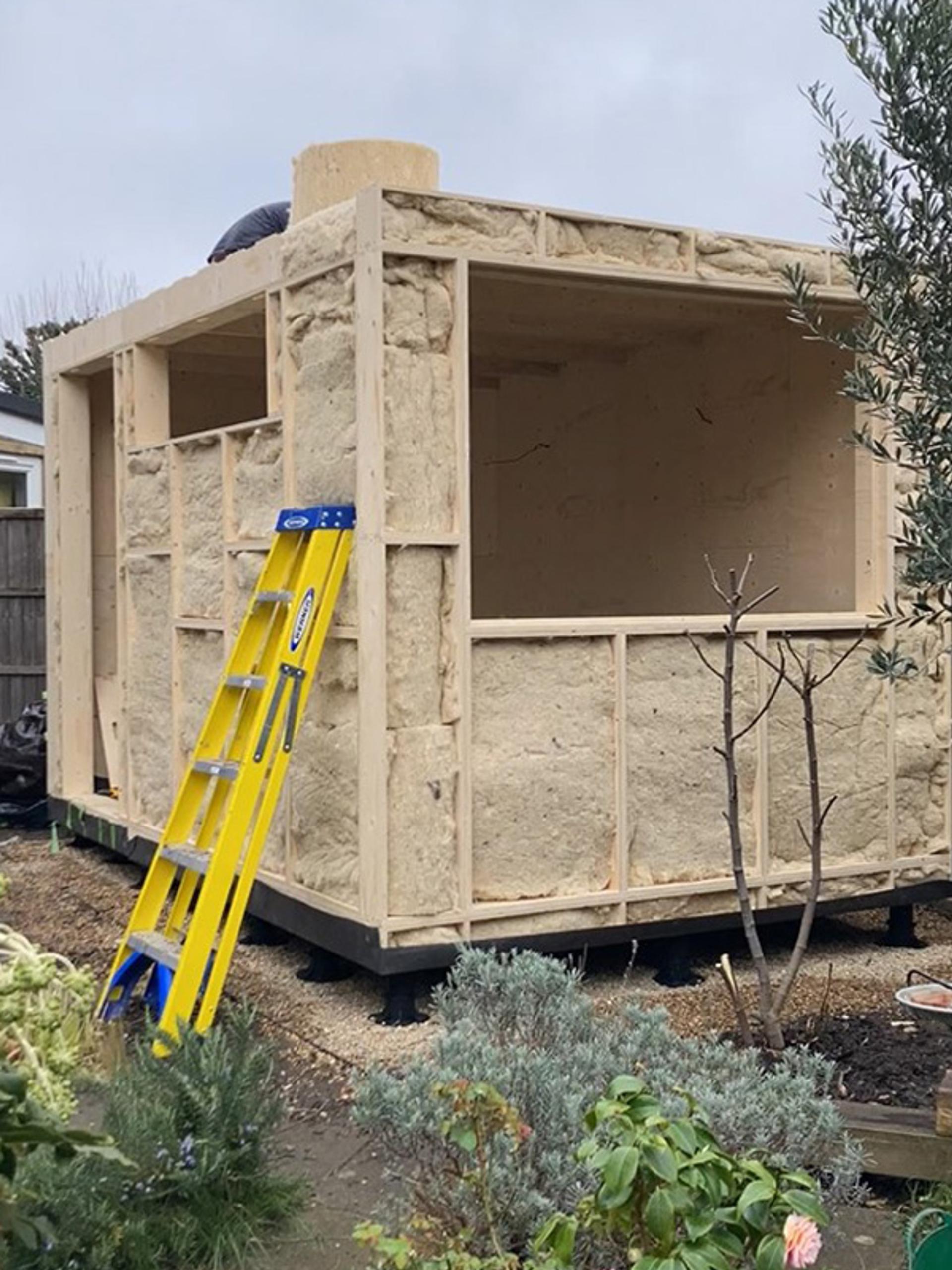 A partially built timber construction with exposed insulation panels, a yellow ladder leaning against it, and plants and shrubs surrounding the sustainable construction area.
