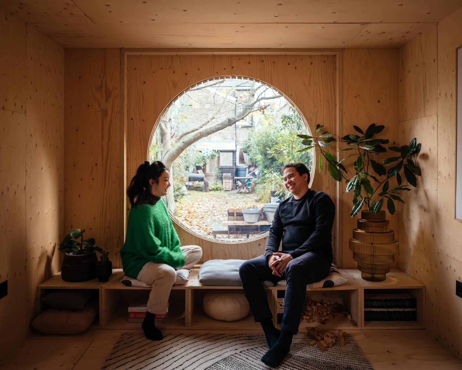 Two people sit and talk in a cosy birch-panelled room with a large round window overlooking a garden. Indoor plants and cushions add to the warm, inviting atmosphere created by timber construction.