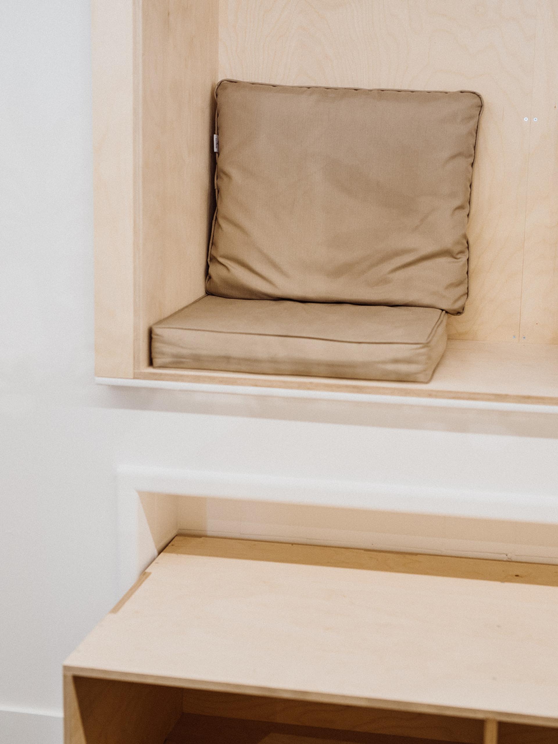 A built-in wooden nook crafted from spruce with a beige seat cushion and matching back cushion. The light-coloured timber construction adds warmth, whilst an open storage space sits conveniently below the nook.