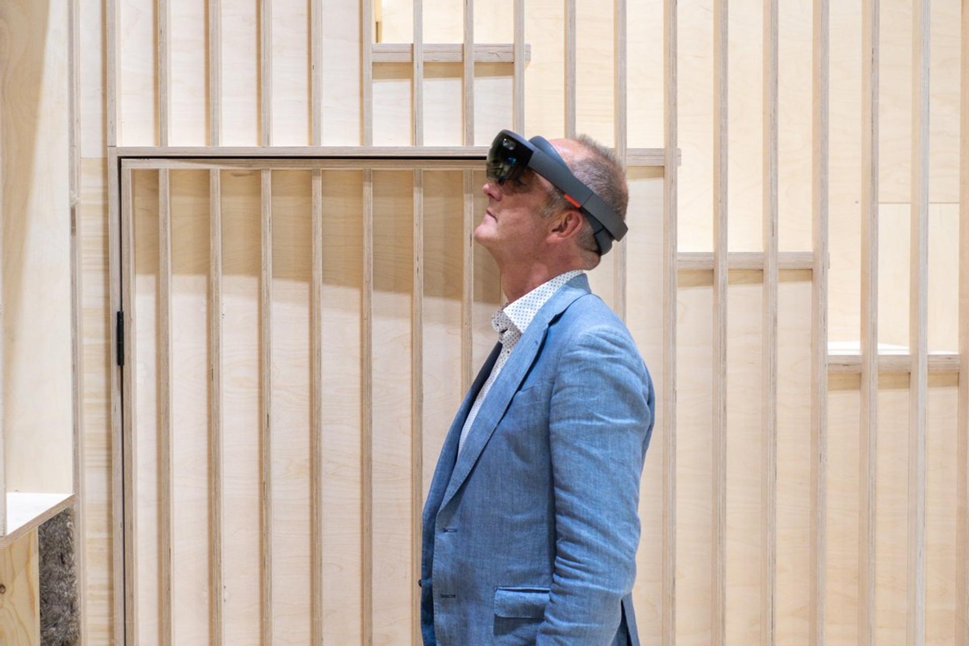 A man in a blue blazer and virtual reality headset looks upwards whilst standing in front of a sustainable light birch slatted wall.