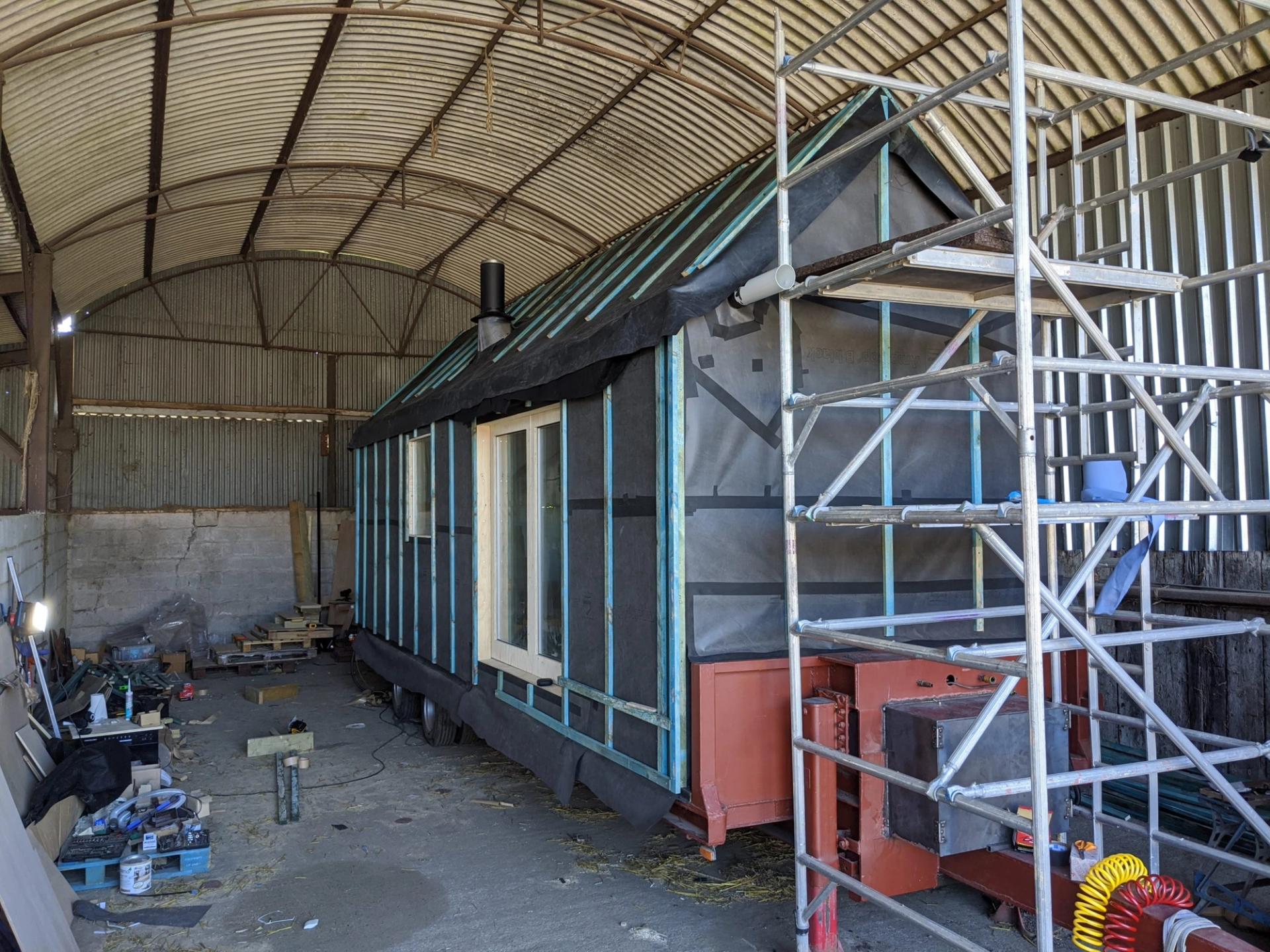 A partially constructed modular tiny house on wheels sits inside a large corrugated metal barn. Scaffolding stands beside the house, and various sustainable construction materials and tools are scattered around.