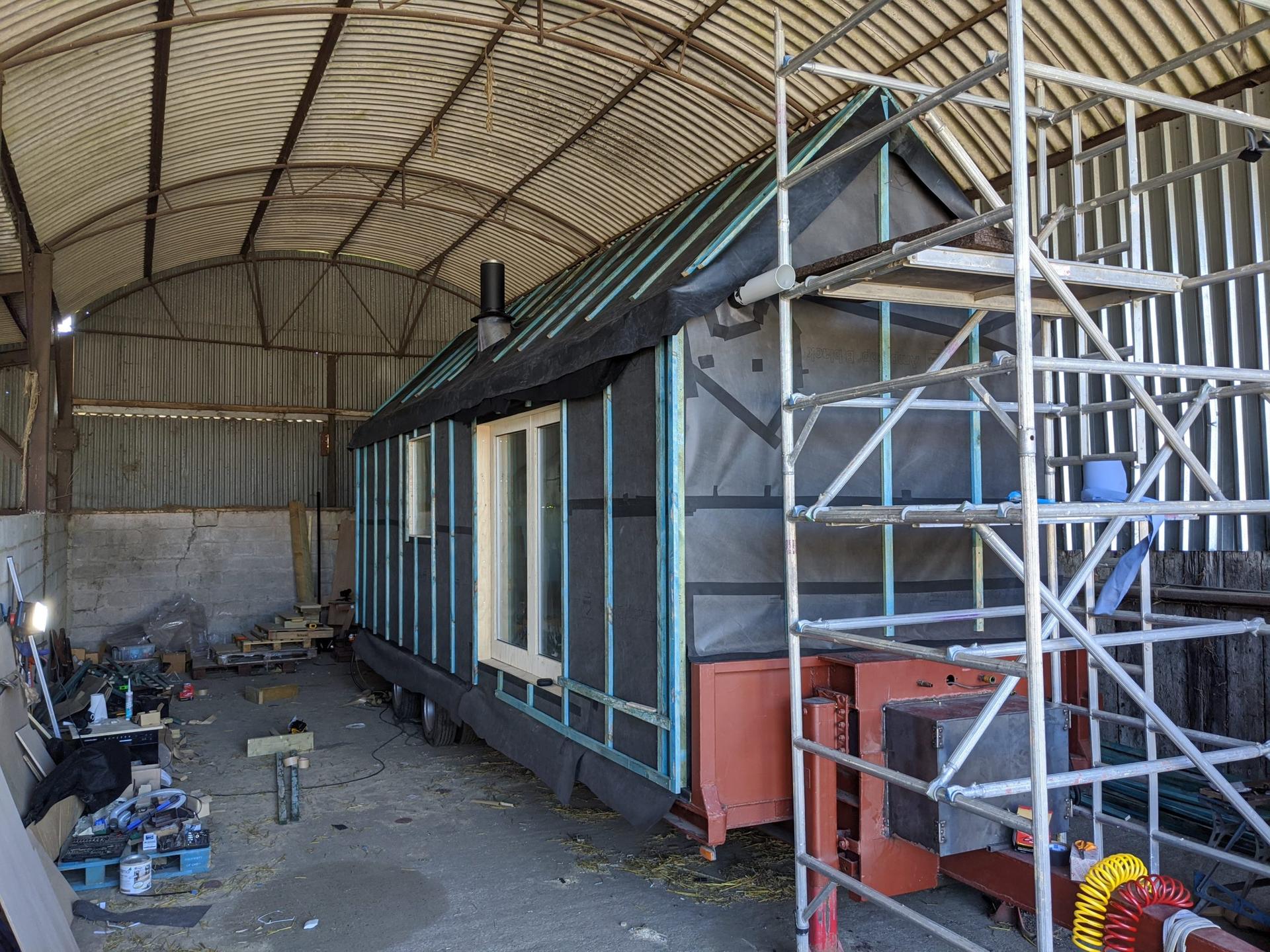 A partially constructed modular tiny house on wheels sits inside a large corrugated metal barn. Scaffolding stands beside the house, and various sustainable construction materials and tools are scattered around.