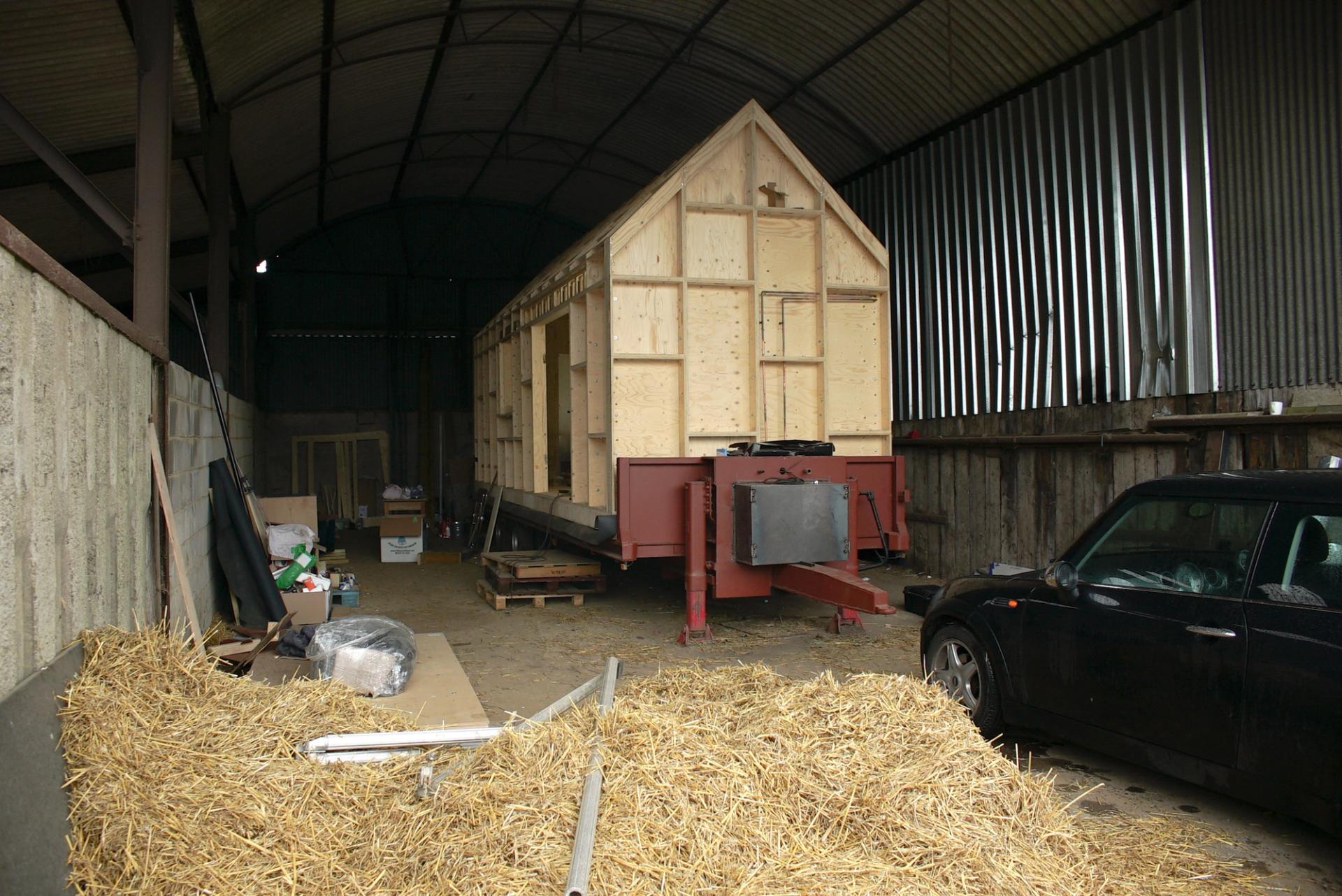 A partially constructed modular tiny house on a trailer sits in a large shed. Nearby are a black car, bales of straw, and scattered tools. Corrugated metal walls and a high arched roof enclose the space. Spruce beams accent the construction.