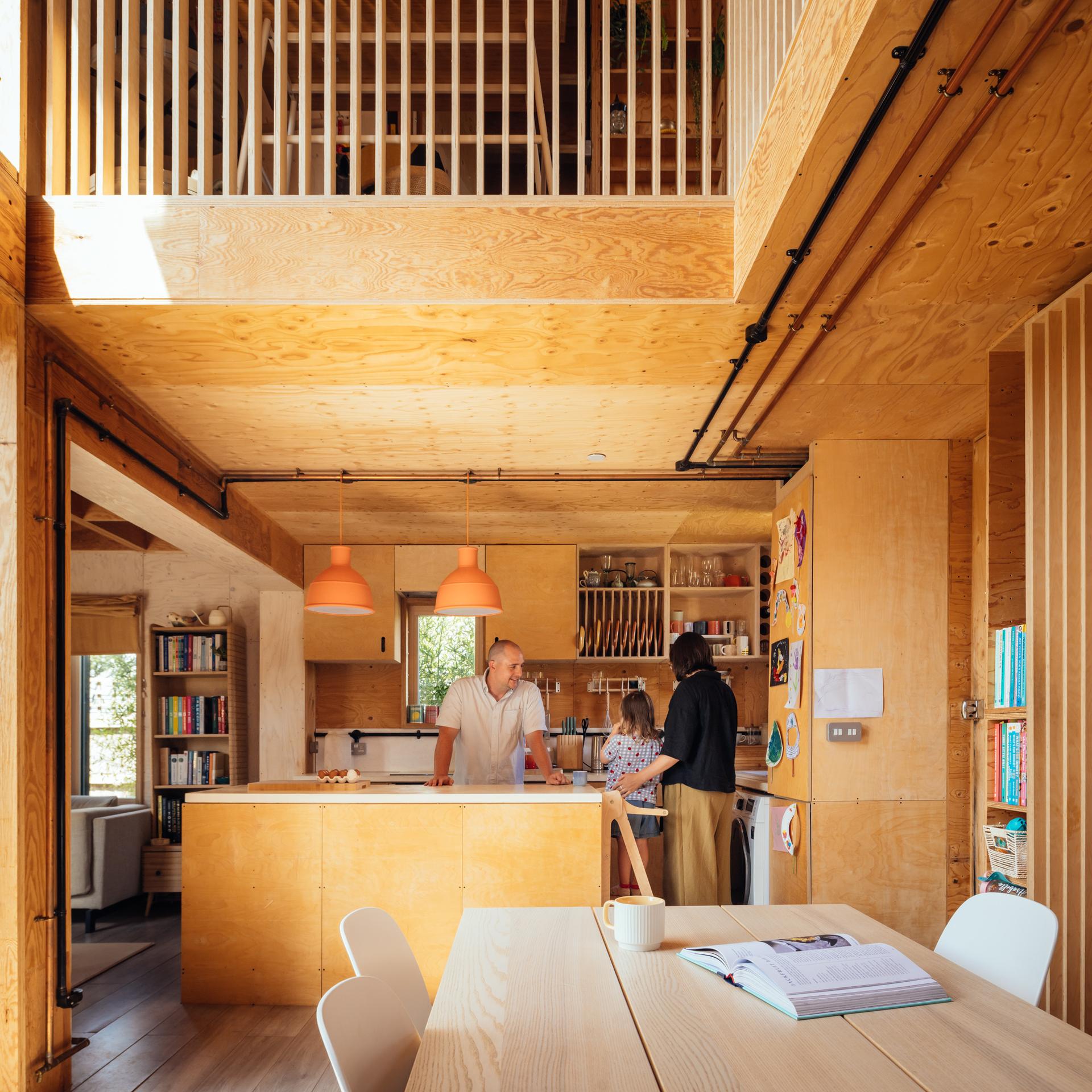 A family stands in a light wood kitchen featuring a box house extension, an island, two pendant lights, and a mezzanine above.