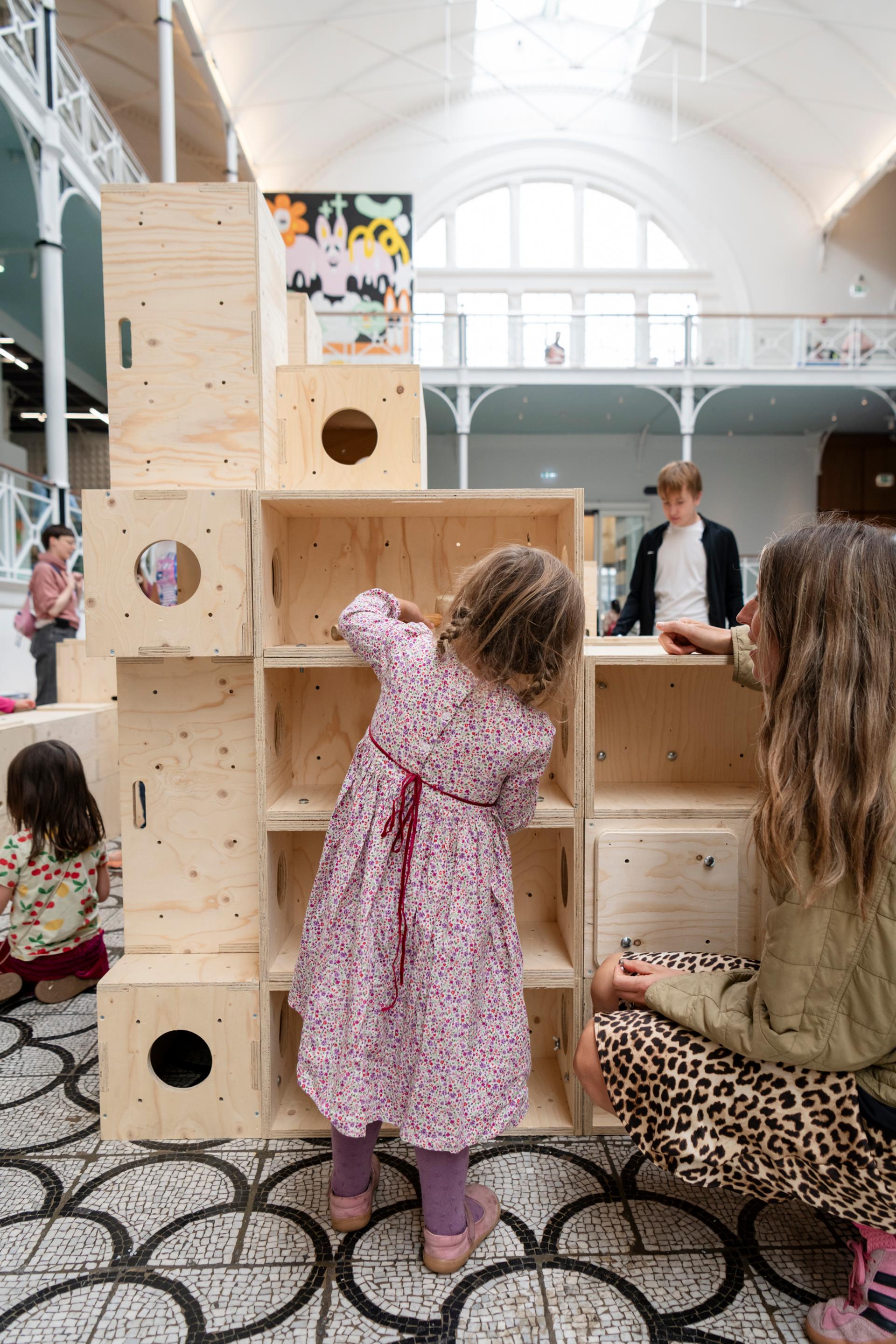 A young girl in a floral dress plays with large U-Build wooden blocks indoors, while an adult and other children are nearby. The bright space features a high skylight and a patterned tiled floor.