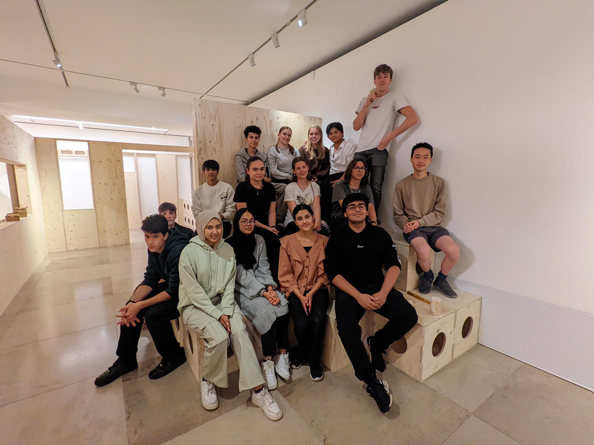 A group of sixteen young adults sit and stand on modular wooden benches in a modern, minimalist room with white walls and a polished floor, posing and smiling for a group photo.