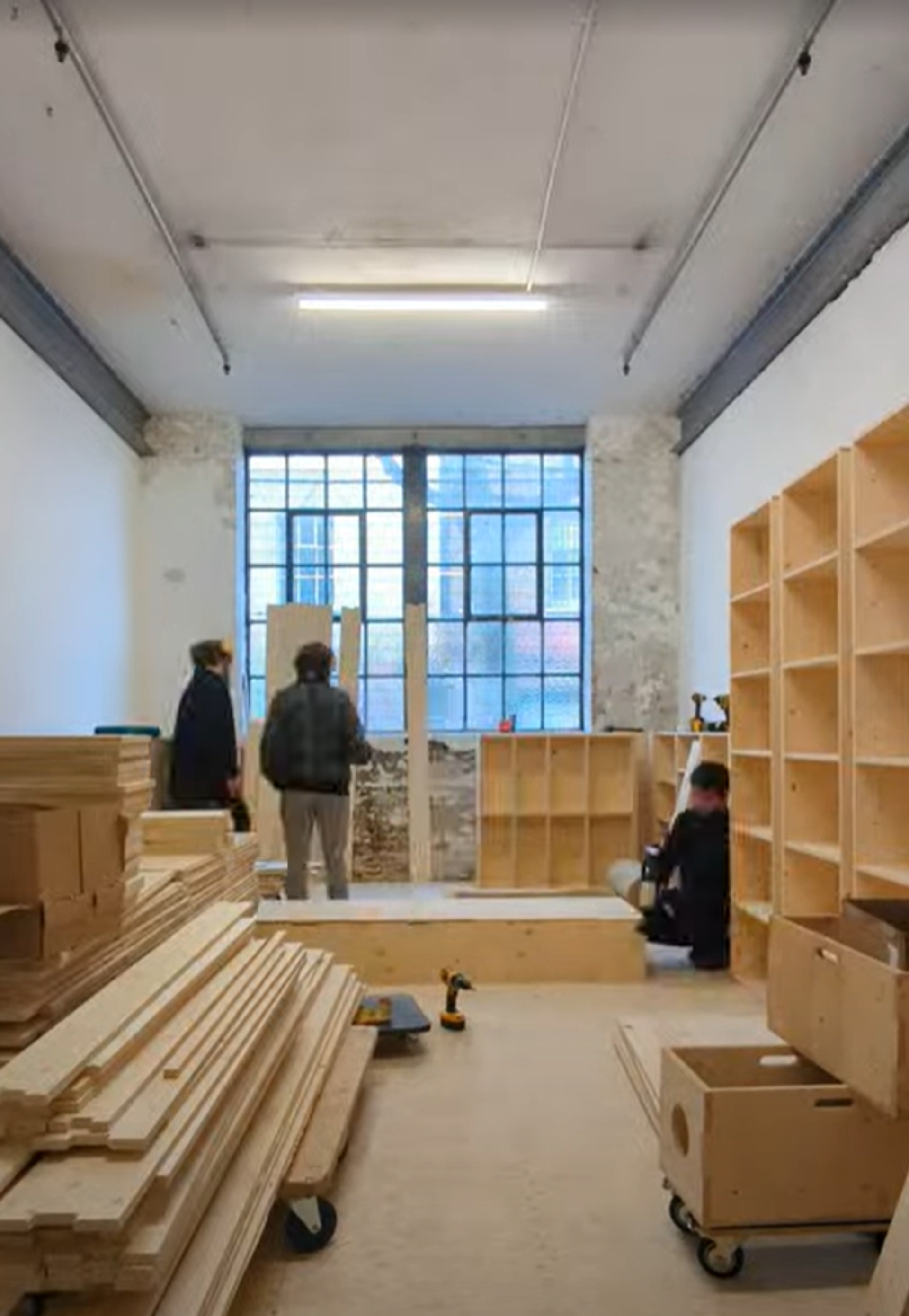 Three people are assembling sustainable spruce shelves in a room with high ceilings, a large window, and exposed brick. Wooden planks and boxes are stacked on the floor as they organise the shelves along the walls.