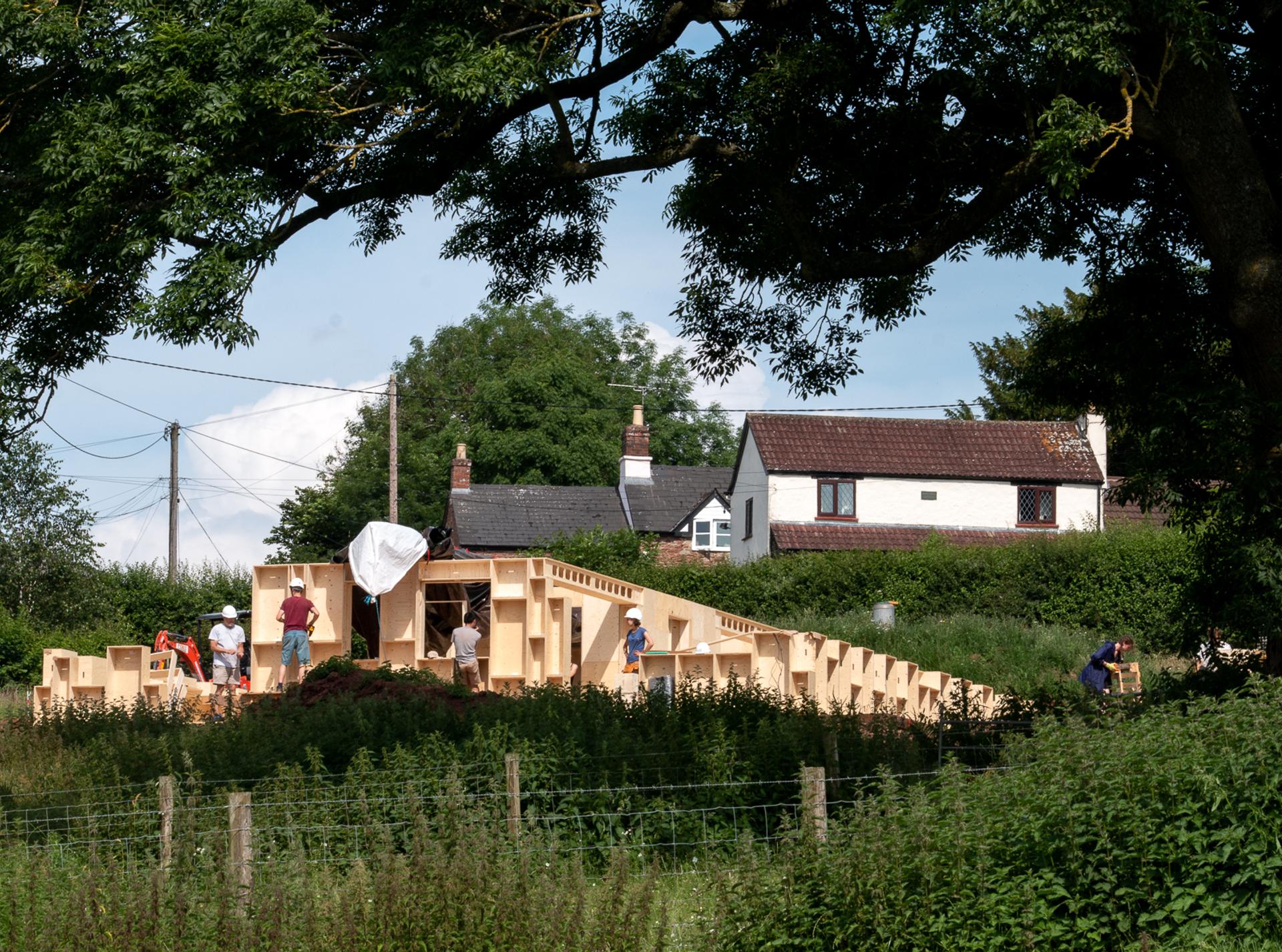 A group of people work on a wooden structure with geometric shapes in a grassy area, embracing reuse and sustainability, with houses and trees in the background on a sunny day.