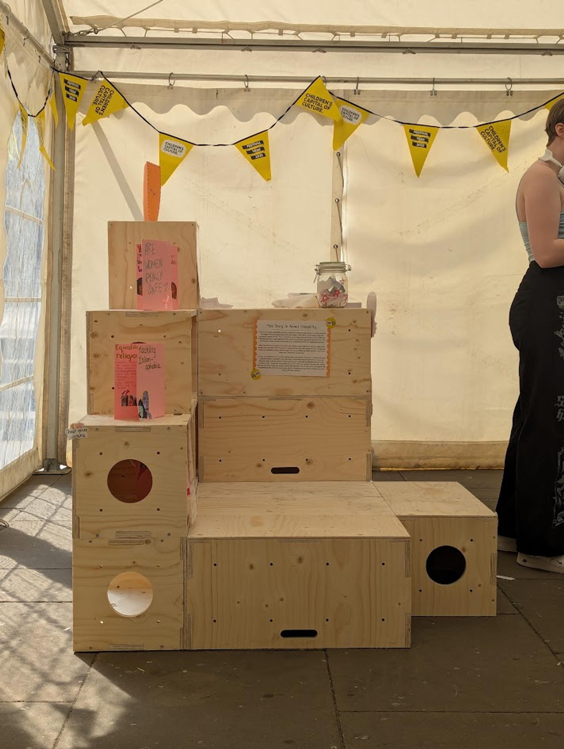 Wooden boxes made from birch plywood are stacked to form steps inside a marquee, decorated with yellow bunting. Books and a jar sit on top, and a person stands partially out of shot on the right, highlighting the sustainable set-up.