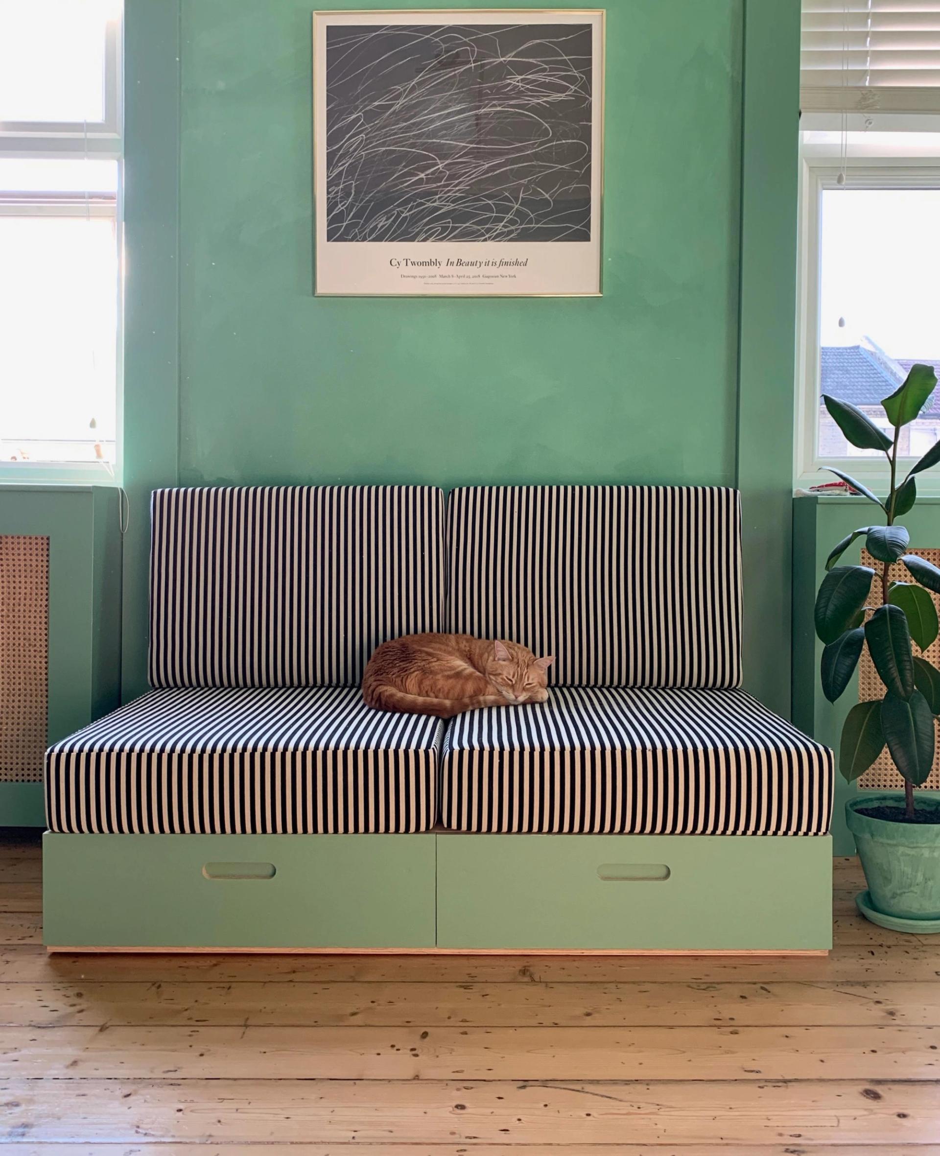 A ginger cat sleeps curled up on a black-and-white striped upcycled sofa in a room with mint green walls and wooden floors. A potted plant and framed artwork decorate the space.