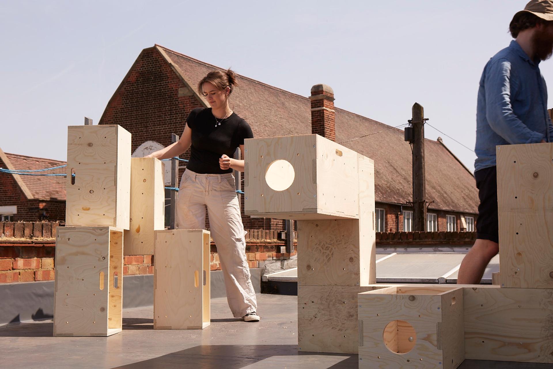 Two people assemble modular plywood blocks with cut-out holes on a rooftop, framed by brick buildings in the background on a sunny day.