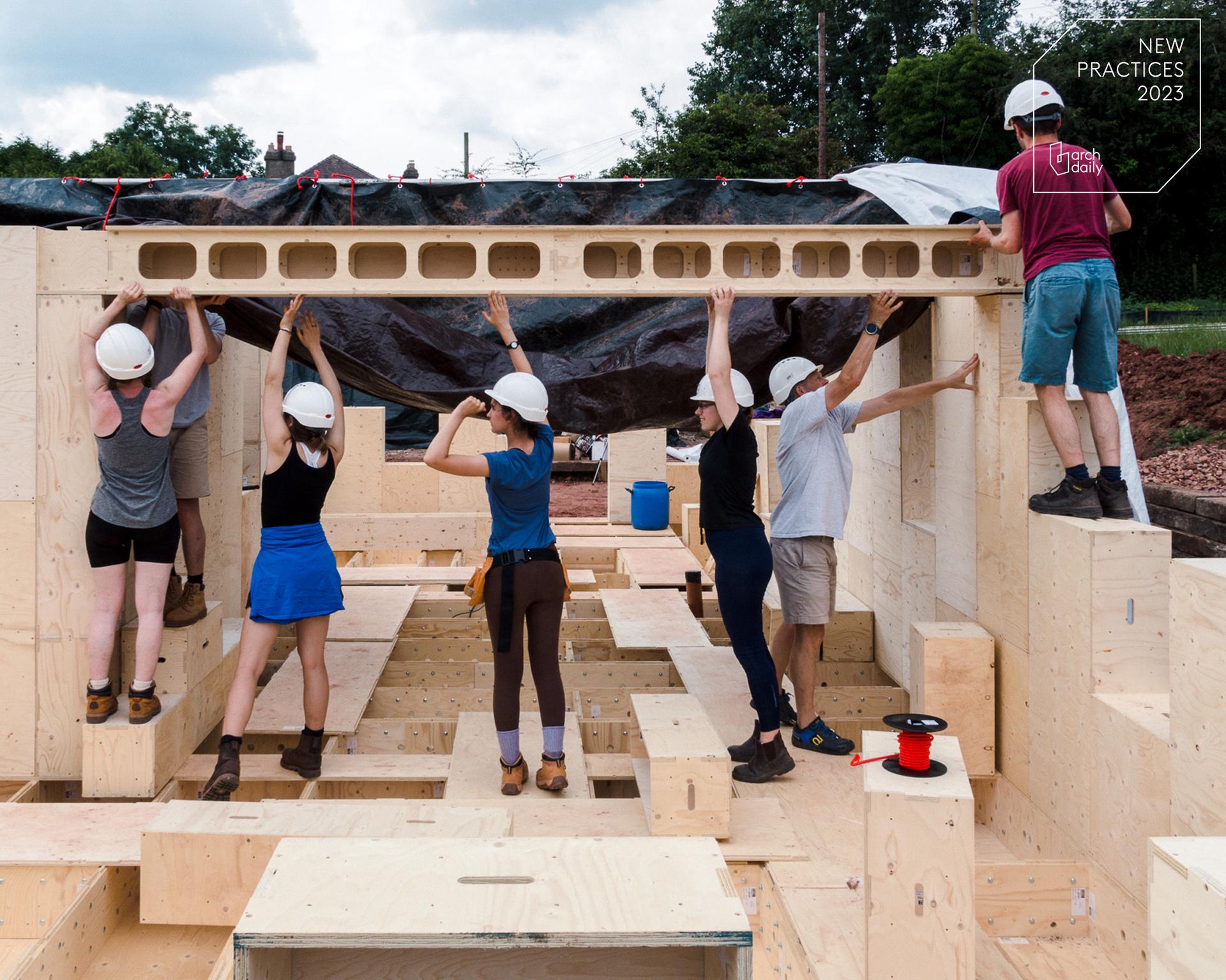 Six people wearing hard hats work together to lift and position a large spruce beam on a building site made of light-coloured timber, outdoors, with trees in the background.