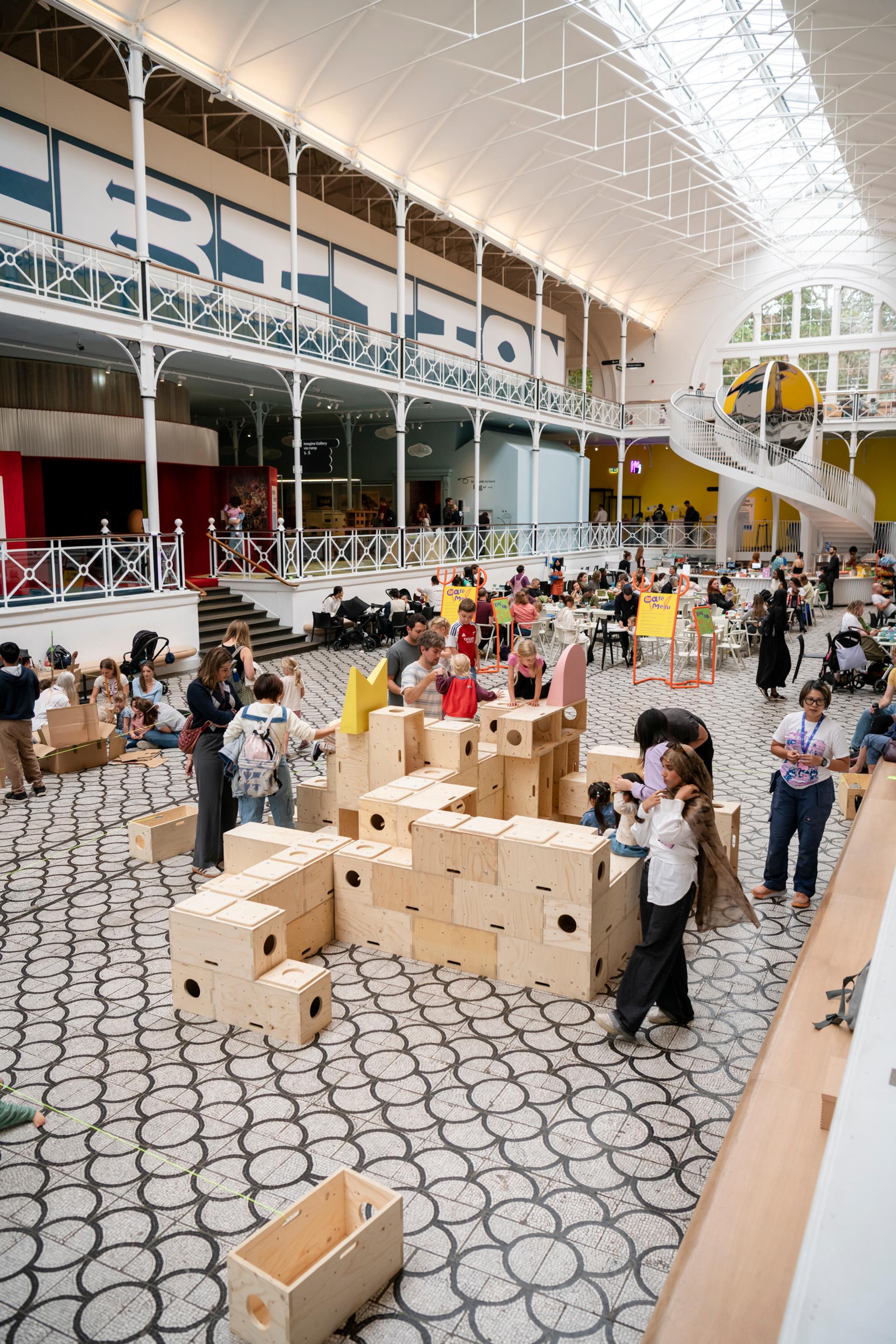 Children and adults play and build with large U-Build wooden blocks in a bright, spacious indoor hall with patterned flooring and a high, arched glass ceiling. Other visitors observe and walk around in the lively, interactive space.