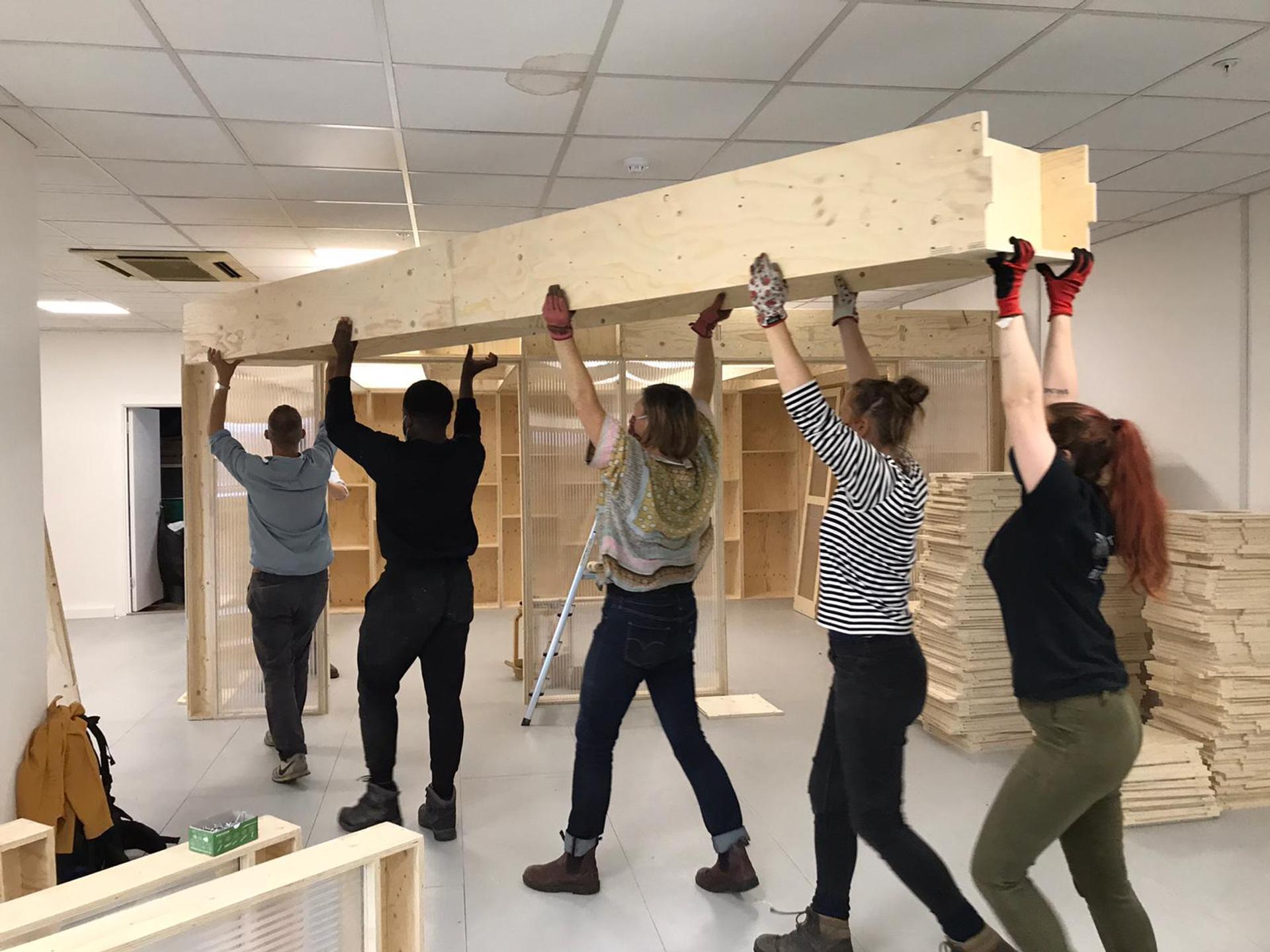 Five people work together to lift and position a large spruce beam, highlighting sustainable timber construction, inside a room with shelves and stacks of timber panels in the background.